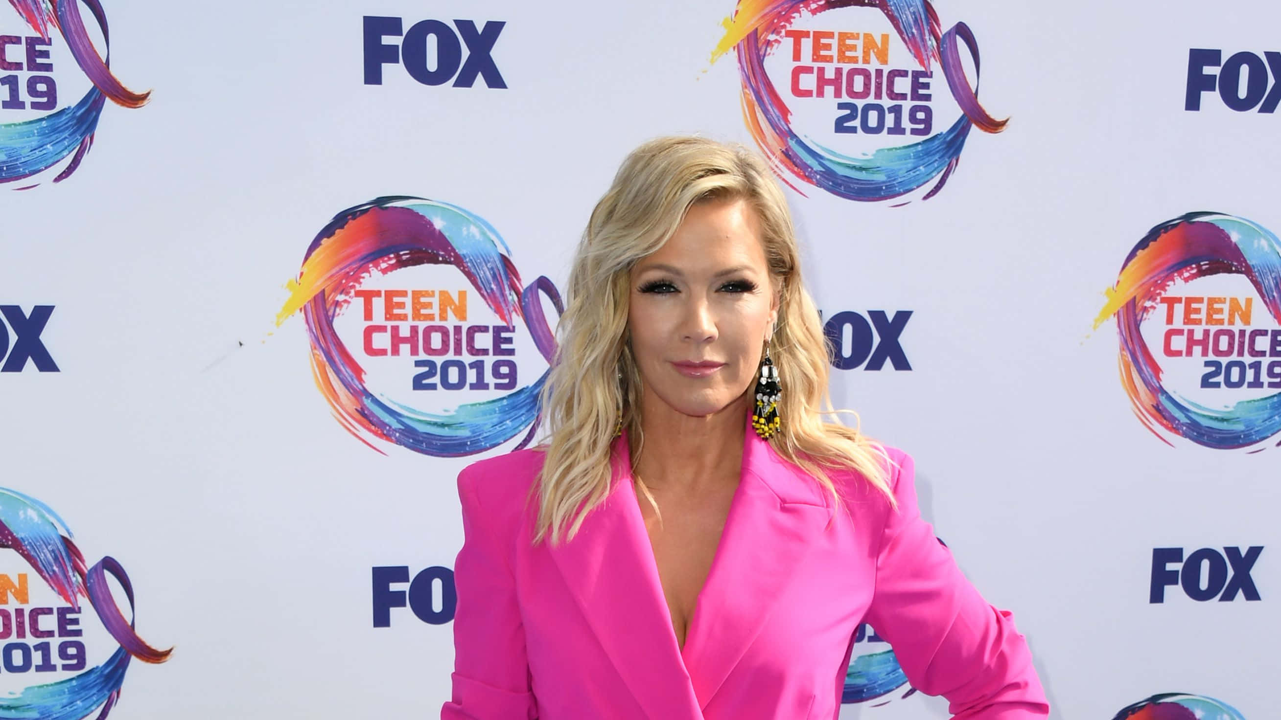 Jennie Garth 桌布 壁紙 — 一位金髮女性穿著亮粉紅色西裝外套，在Teen Choice 2019紅地毯上自信地擺姿勢，背景帶有品牌標誌。