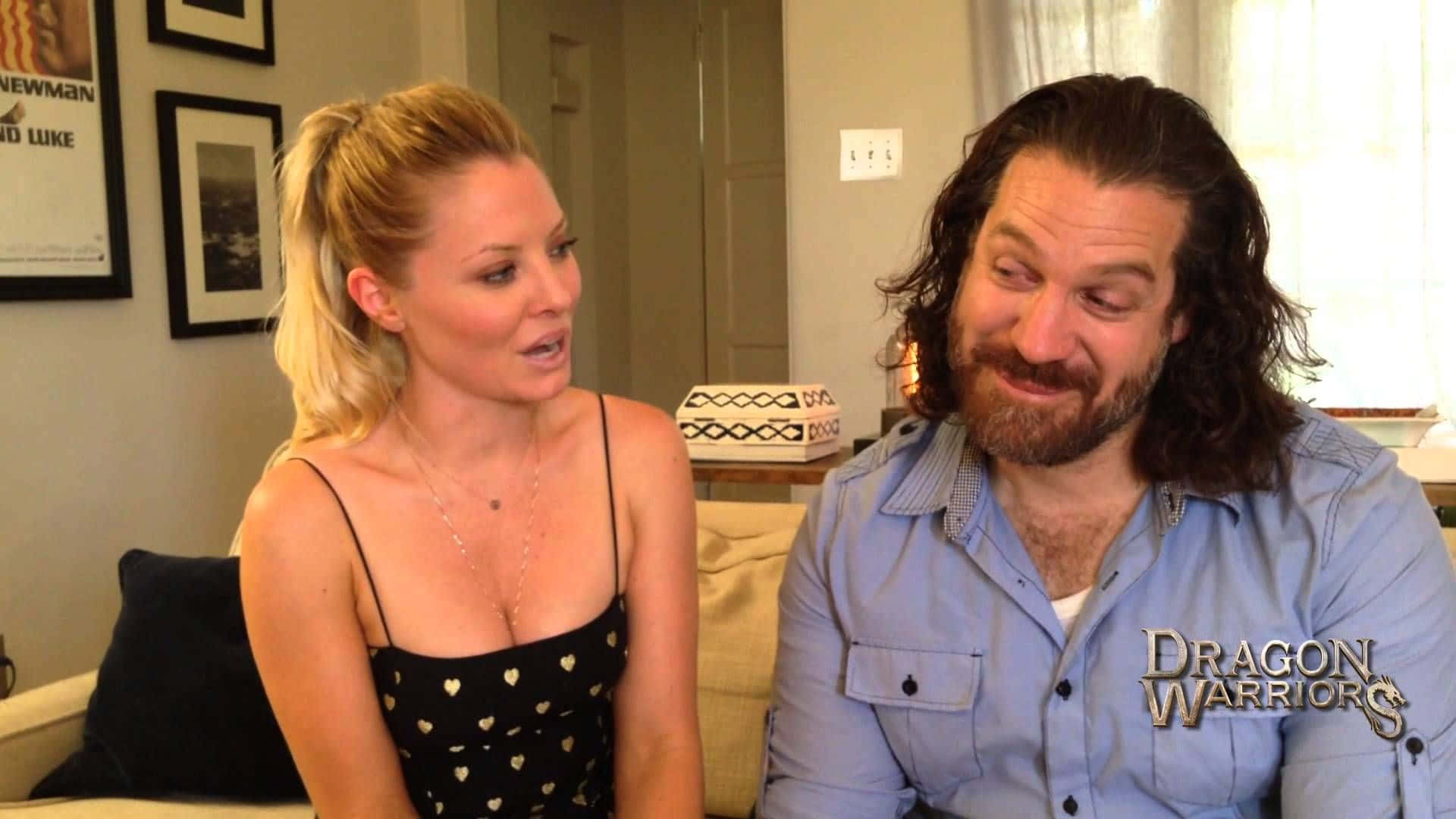 Kaitlin Doubleday 桌布 壁紙 — 一位金髮女子身穿黑色心形圖案上衣，一位留鬍鬚的男子身穿藍色襯衫，坐在梳化上，在燈光溫暖的客廳中交談。