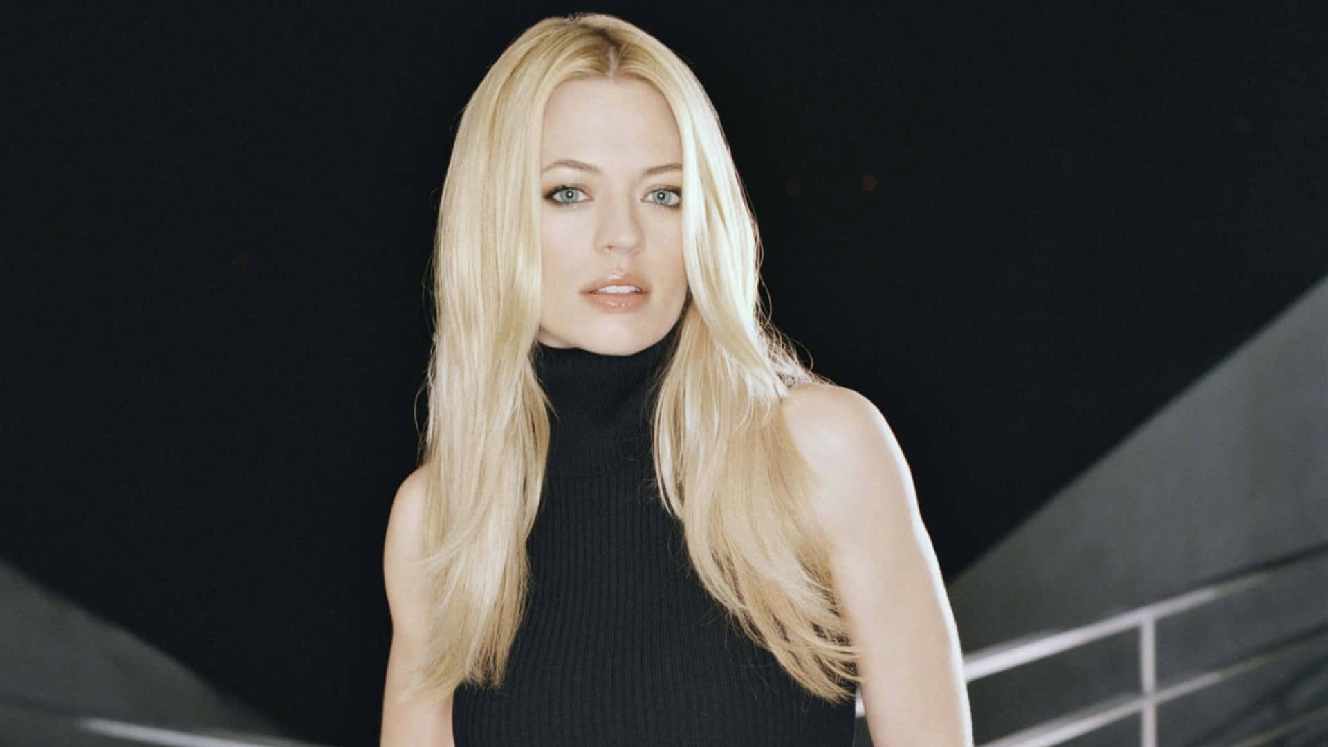 Jeri Ryan 桌布 壁紙 — 一位金髮女子身穿黑色高領毛衣，站在深色幾何背景前，柔和光線突顯她的五官。