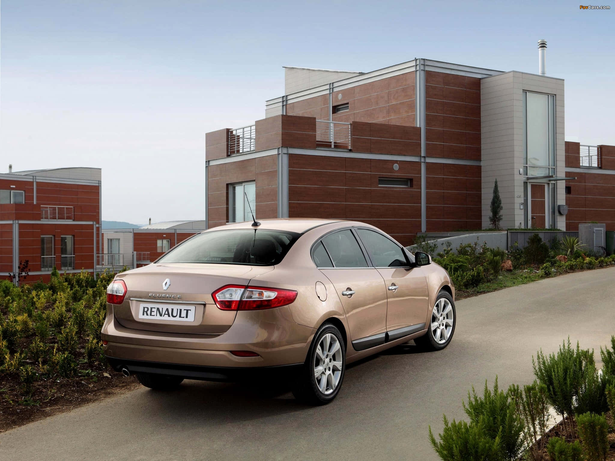 Renault Fluence 桌布 壁紙 — 一輛金色雷諾Laguna房車停泊在鋪砌道路上，旁邊是現代磚砌建築，有綠色灌木，天空晴朗。