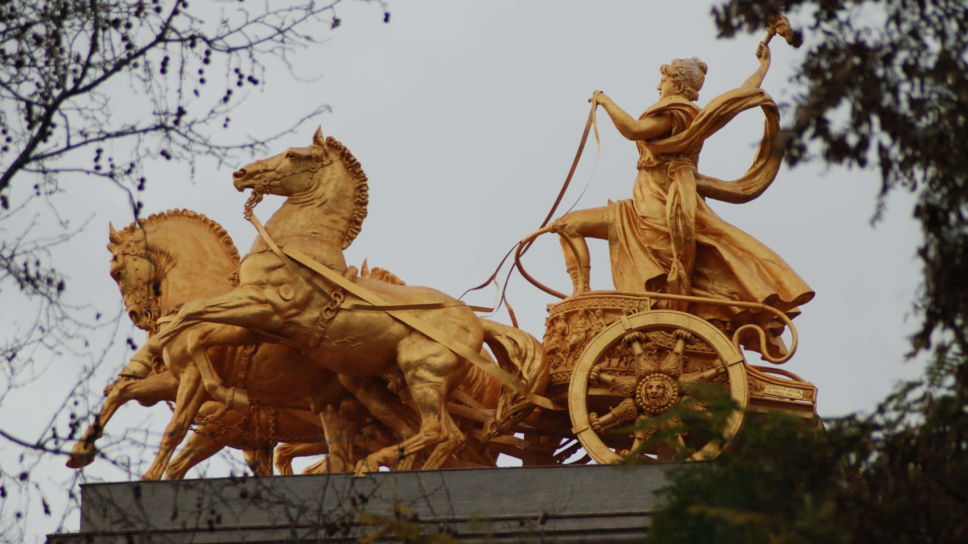  壁紙 — 金色女神像駕駛著由四匹馬拉動的戰車，背景是蒼白的天空，由光禿的樹枝框住。