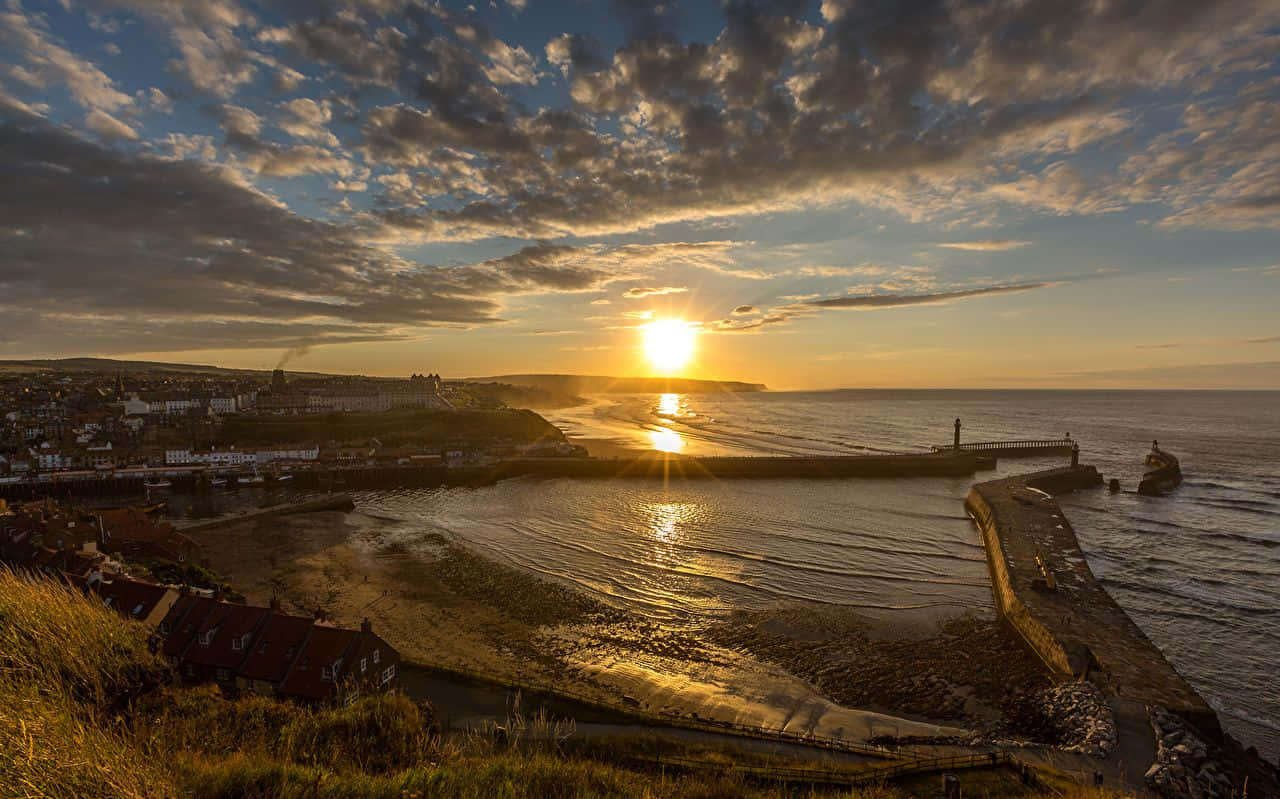 Coastal Sunset 桌布 壁紙 — 金色夕陽照耀著沿海小鎮，石碼頭在戲劇性的雲層下延伸至平靜的水面。