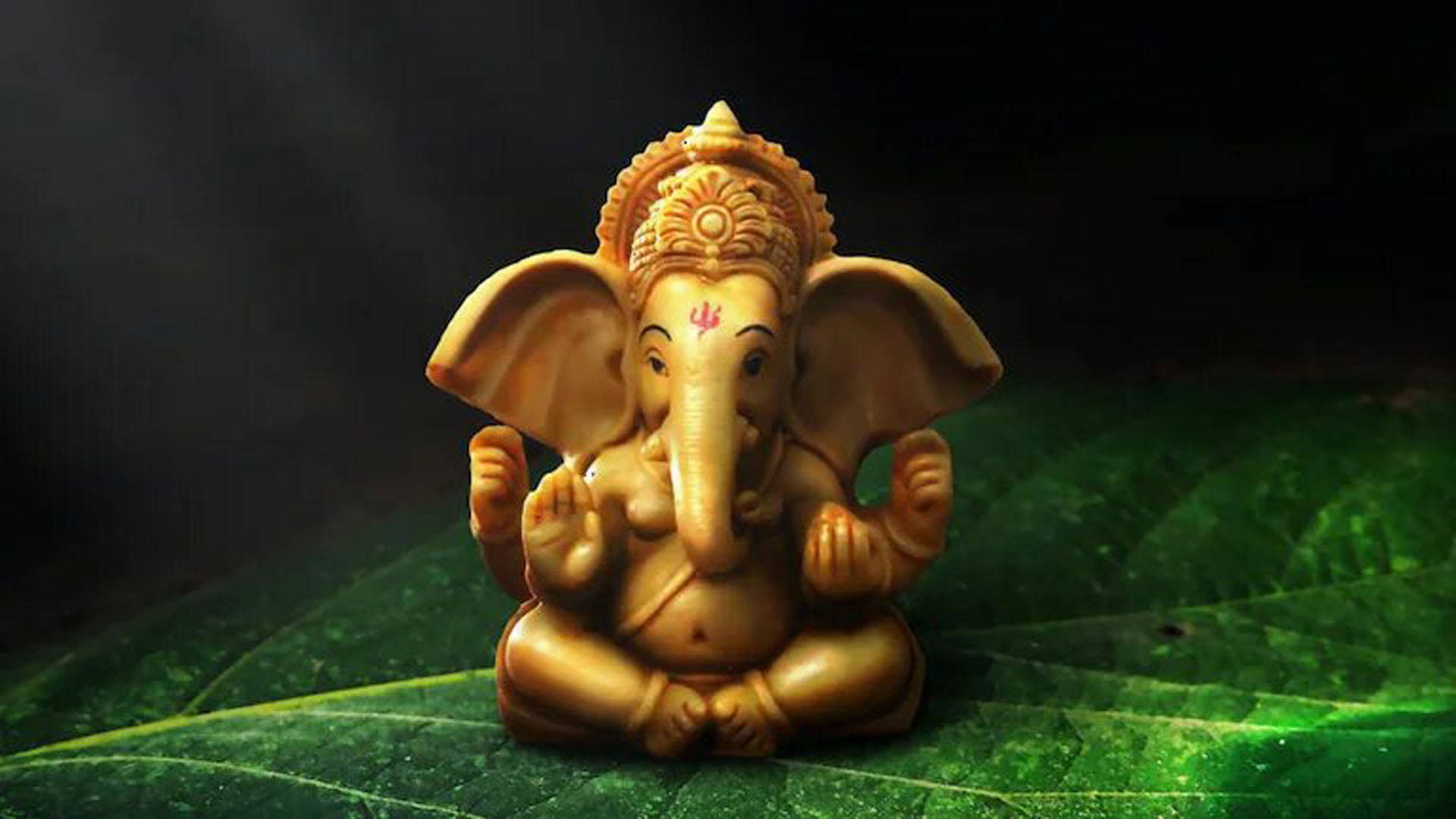 宏觀象徵象神 Ganesh 桌面數位藝術 壁紙 — 金色象神雕像安詳地坐在深色背景的大綠葉上。