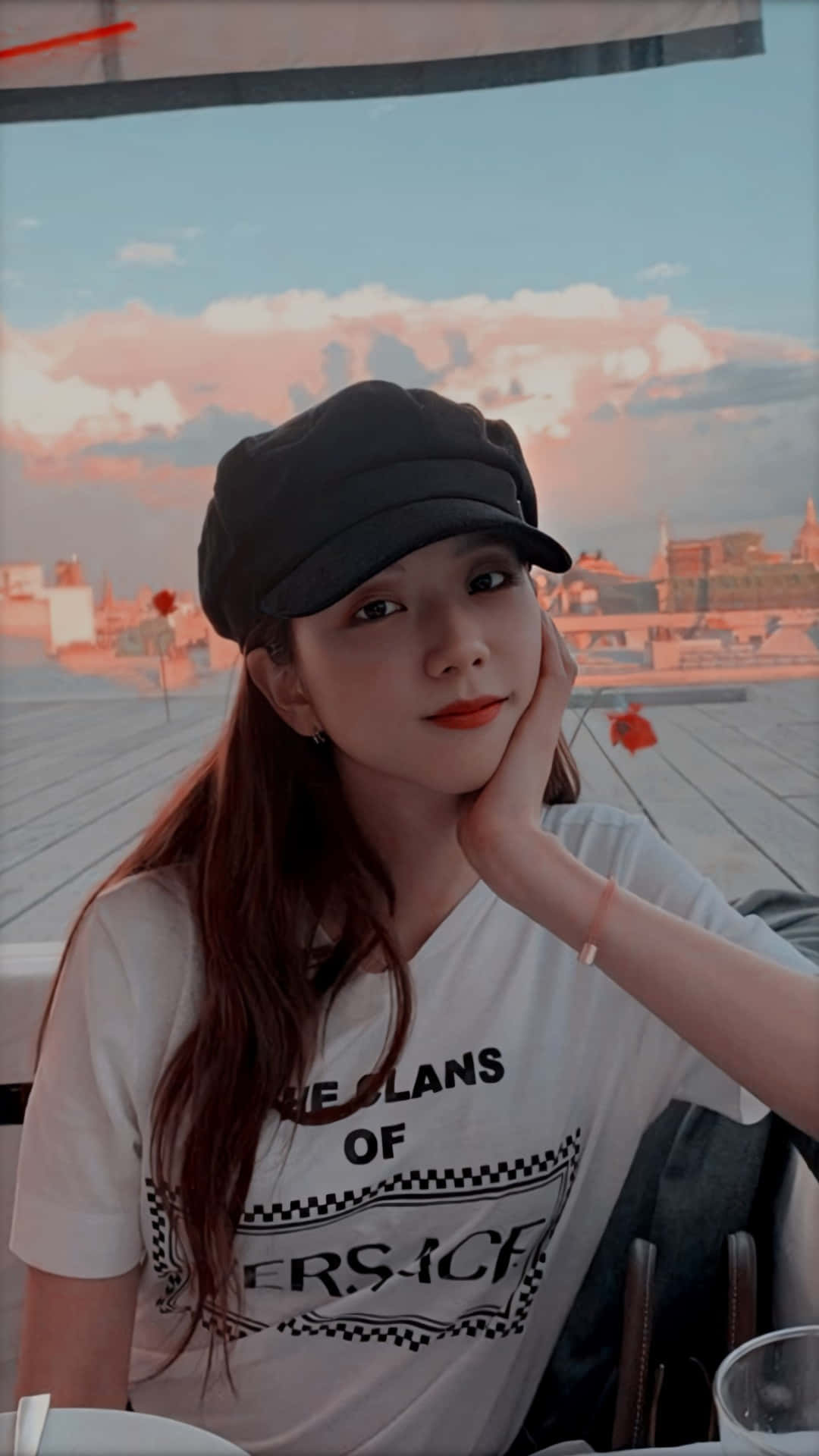 Jisoo BLACKPINK 韓國歌手日常 壁紙 — 一位戴黑色帽子的年輕女子，在夕陽城市景觀背景下，將下巴擱在手上。