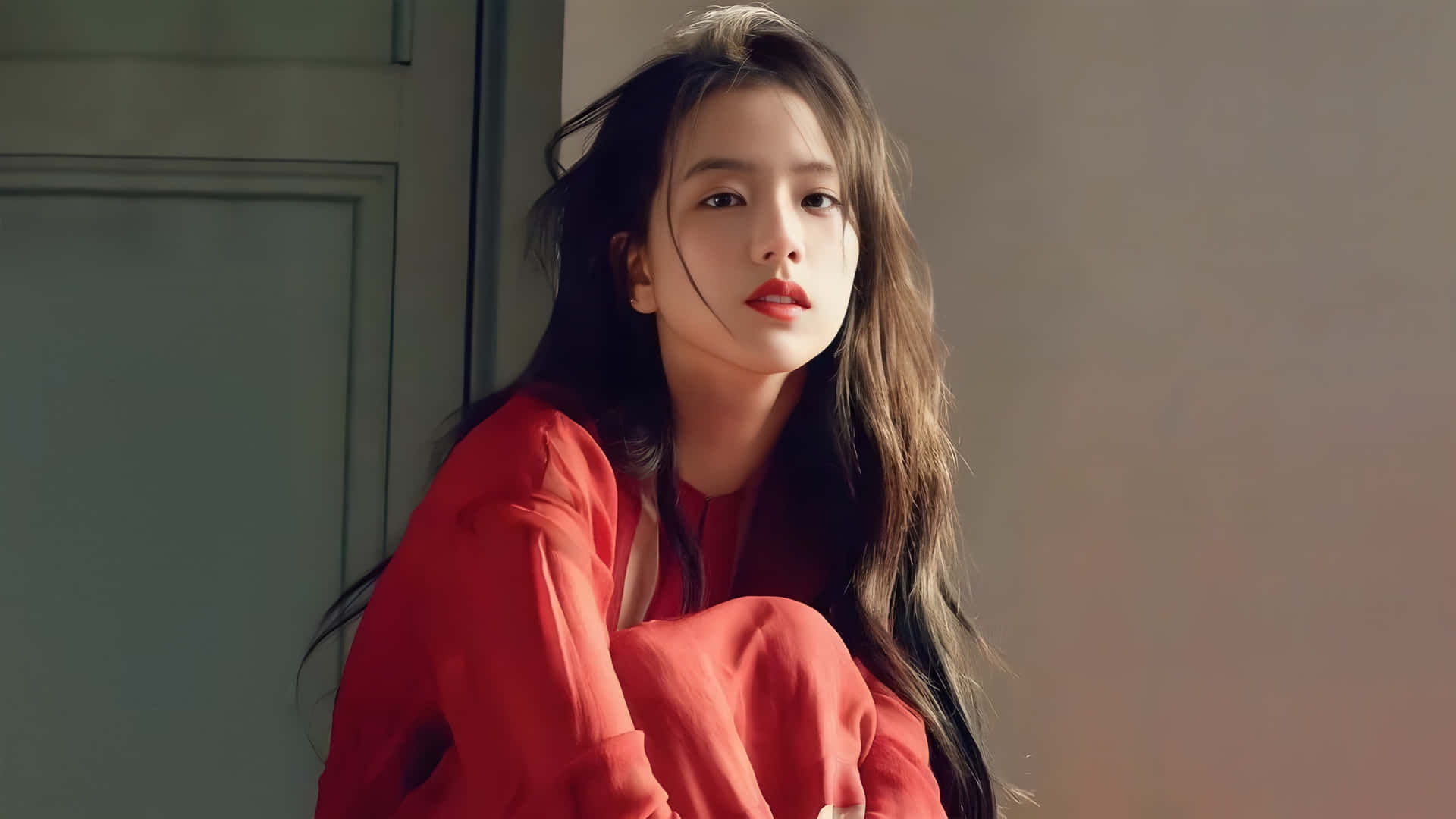 Jisoo BLACKPINK 優雅紅裙造型 壁紙 — 一位身穿紅色襯衫的年輕女子倚在門框上，沐浴在柔和自然光中。