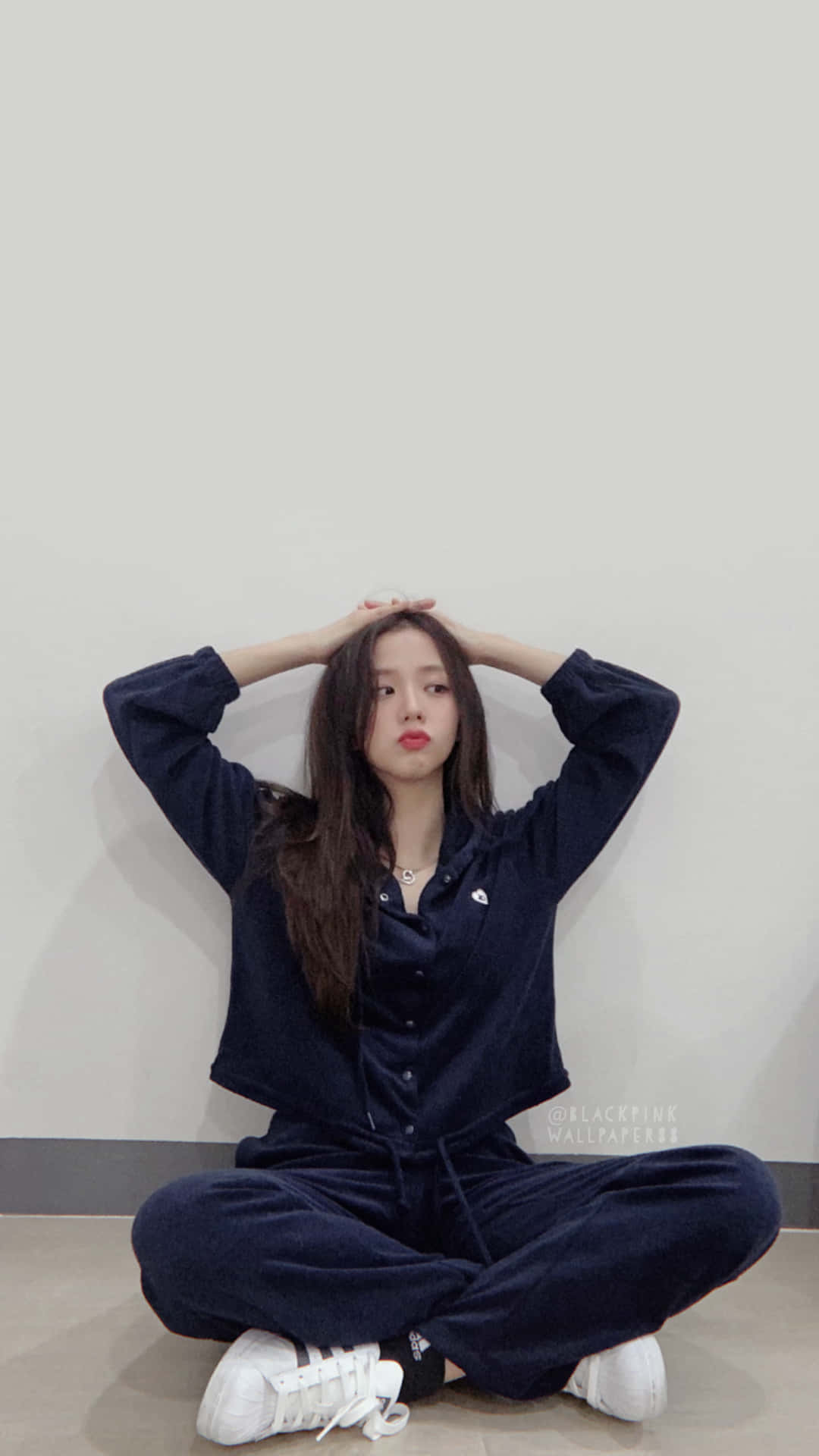 Jisoo BLACKPINK 藍色睡衣造型 壁紙 — 一位年輕女性身穿海軍藍天鵝絨運動服，雙手放在頭上，盤腿坐在淺色地板上，靠著素牆。