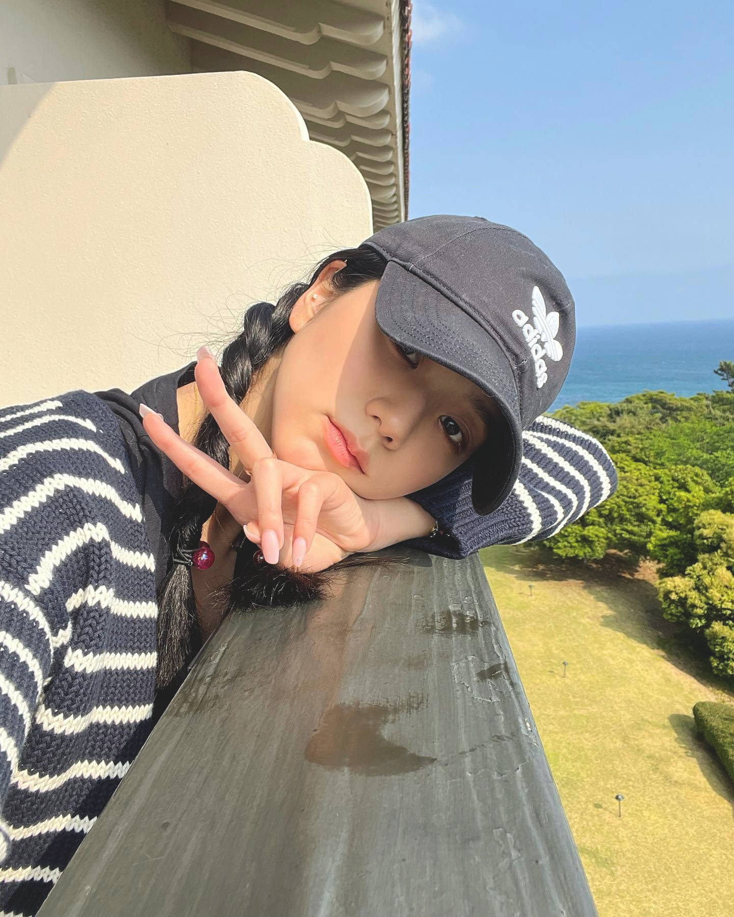 戴帽子和條紋毛衣的年輕女子在陽台上做出和平手勢，俯瞰海洋和綠色山丘。