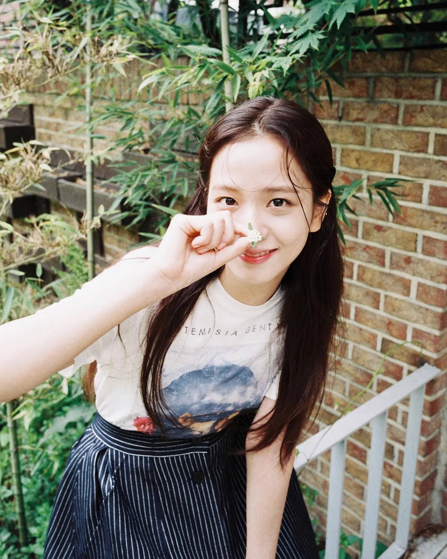 年輕女性將一朵小白花靠近鼻子，在磚牆和竹子附近戶外微笑。