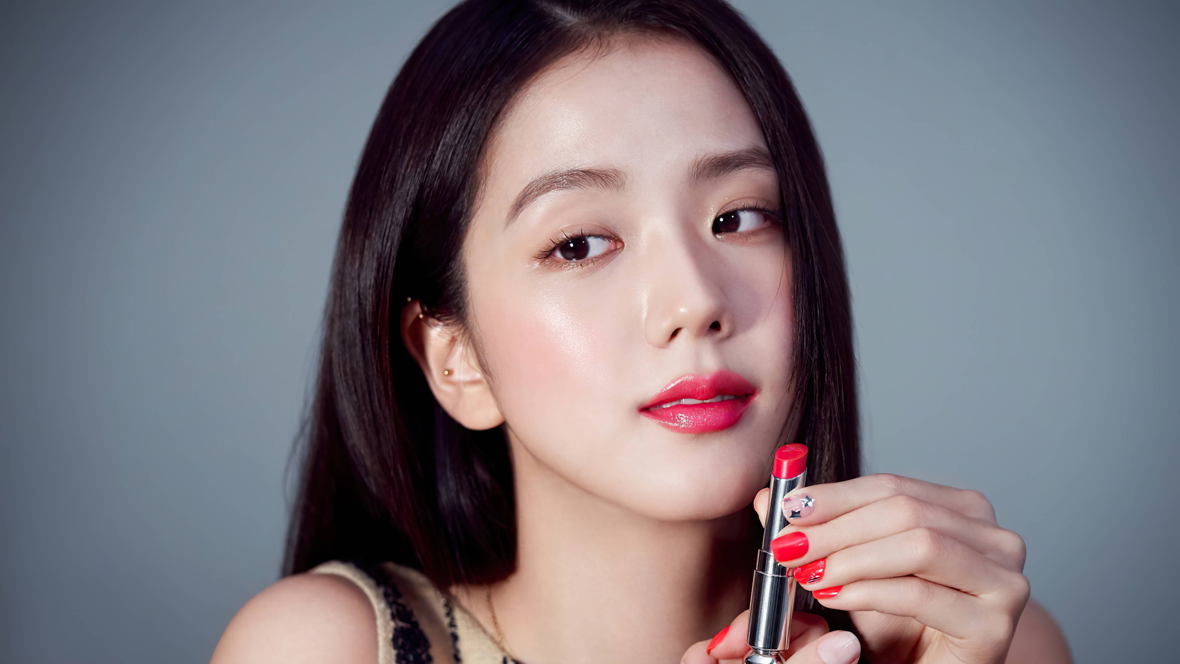 Jisoo 戴迪奧唇膏 壁紙 — 一位深色頭髮、塗著紅色唇膏的年輕女性，在柔和的灰色背景前拿著一支相配的紅色唇膏。