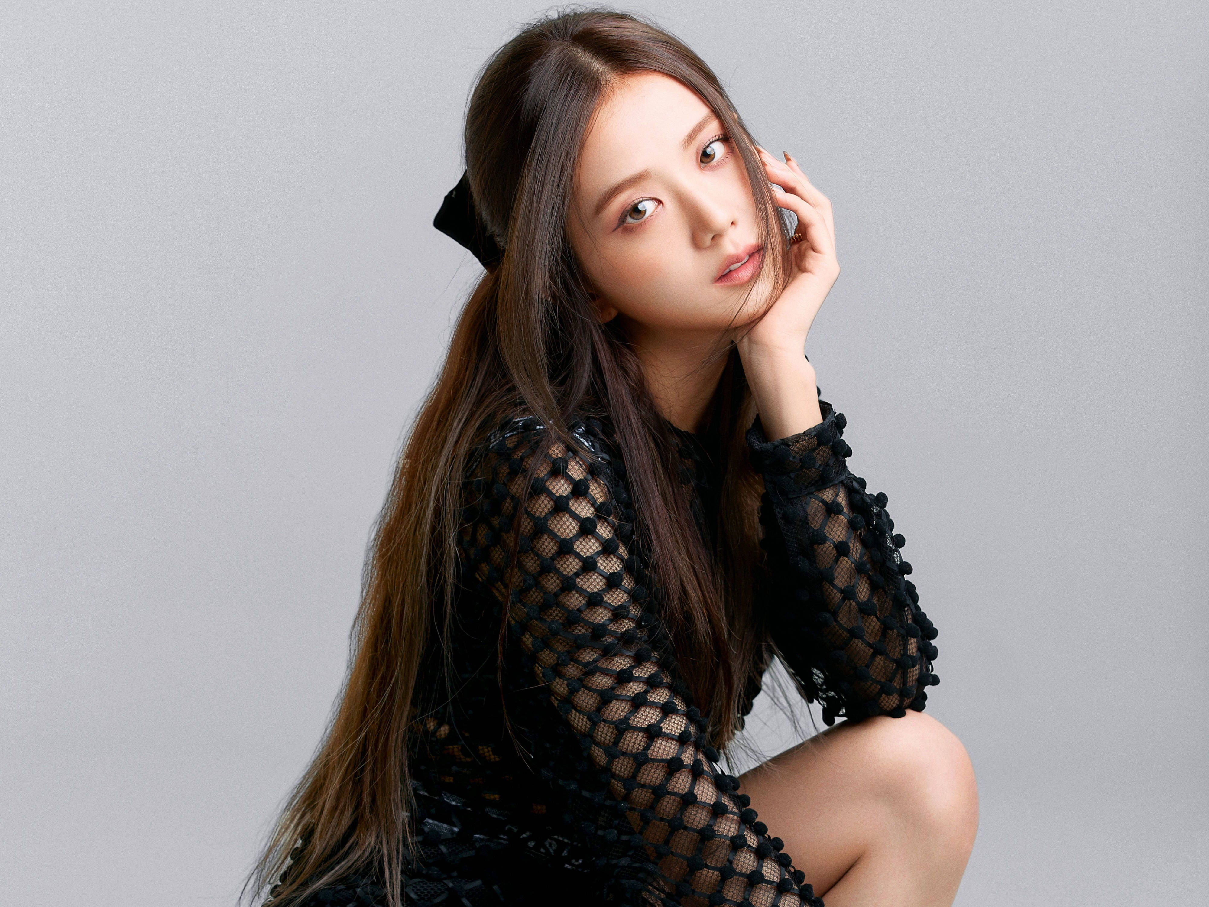 Jisoo 黑色網紗裙 BLACKPINK 桌面壁紙 壁紙 — 一位年輕女性，長長棕色頭髮，戴著黑色蝴蝶結，身穿黑色網狀連衣裙，坐在純灰色背景前，若有所思地凝視著鏡頭。