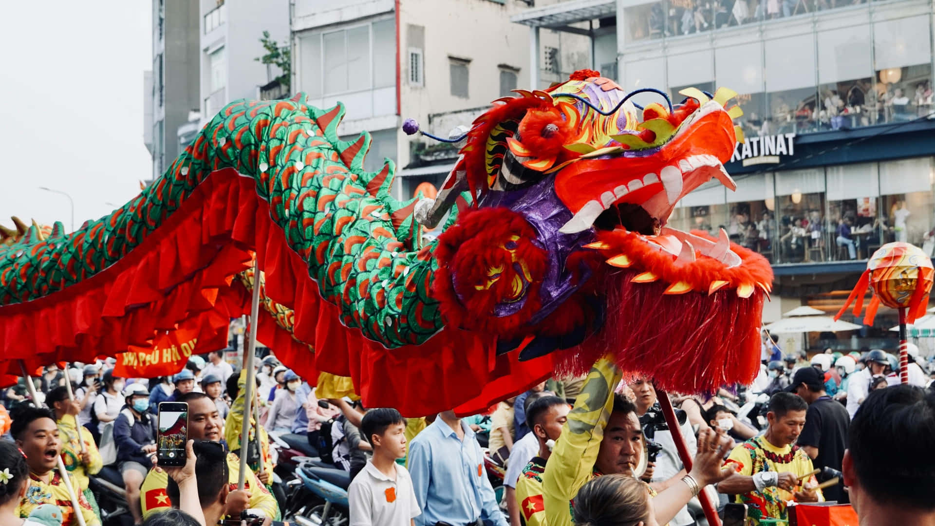  壁紙 — 節日巡遊期間，表演者在擁擠的城市街道上進行充滿活力的紅色和綠色舞龍表演。