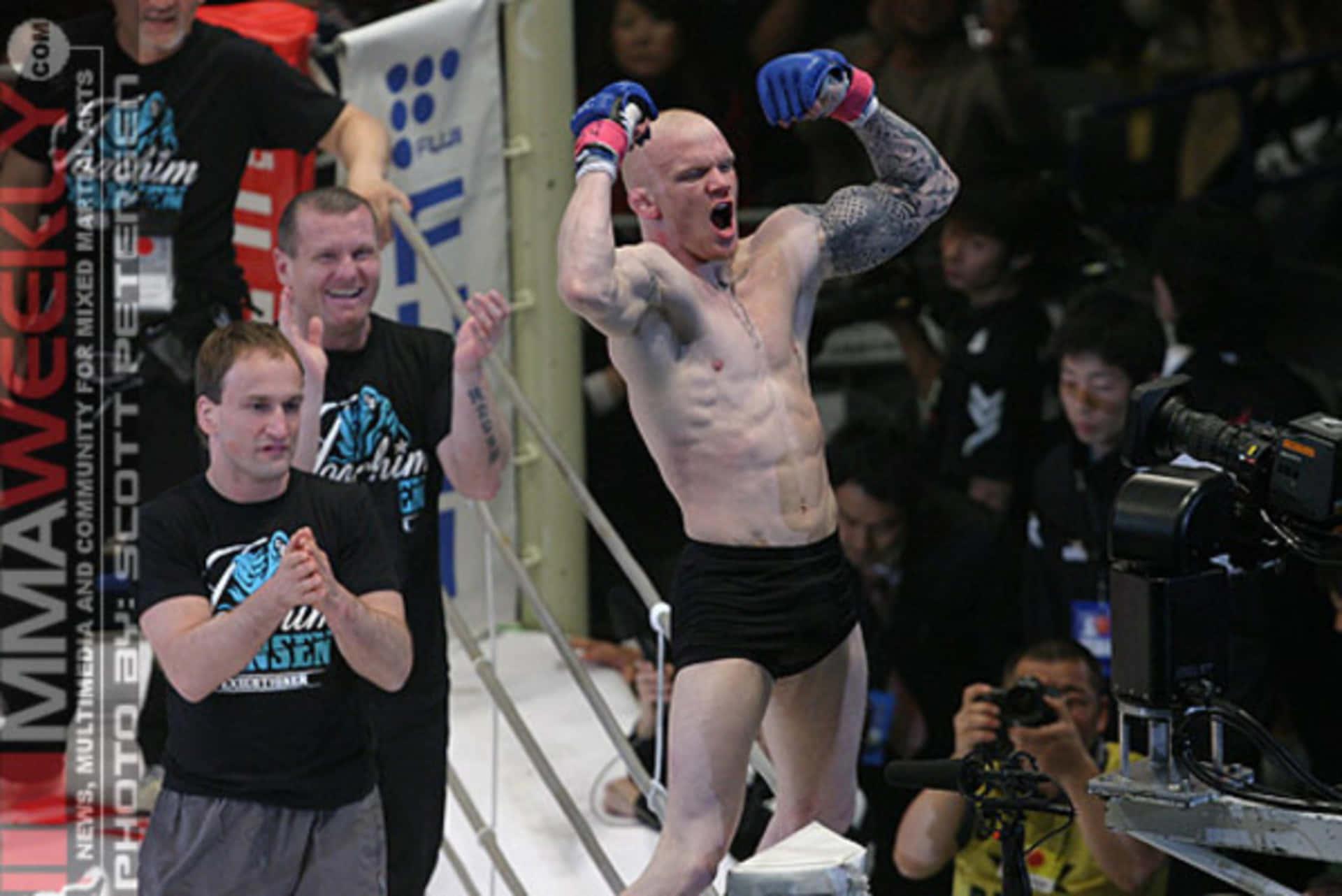 Joachim Hansen 擊敗 Luiz Azeredo 獲勝慶祝 壁紙 — 一位光頭紋身戰士穿著黑色短褲，在MMA八角籠內高舉雙臂勝利咆哮，周圍是粉絲和攝影機。