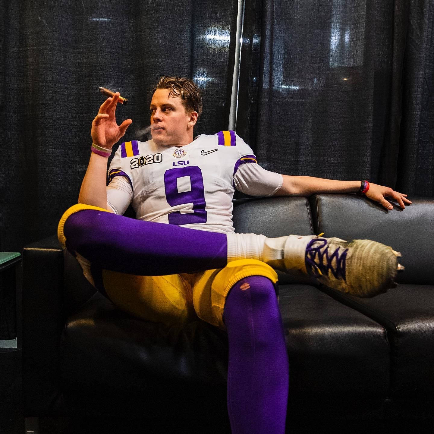 Joe Burrow 吸煙 壁紙 — 一名身穿LSU裝備的美式足球員在黑色沙發上閒躺，手持雪茄，腳上打著石膏。