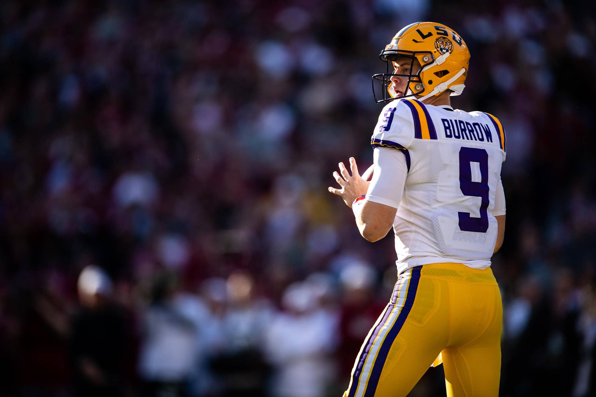 Joe Burrow 黃色頭盔 壁紙 — 身穿LSU球衣的四分衛喬·伯羅在明亮的日間足球比賽中掃視球場，背景是模糊的人群。
