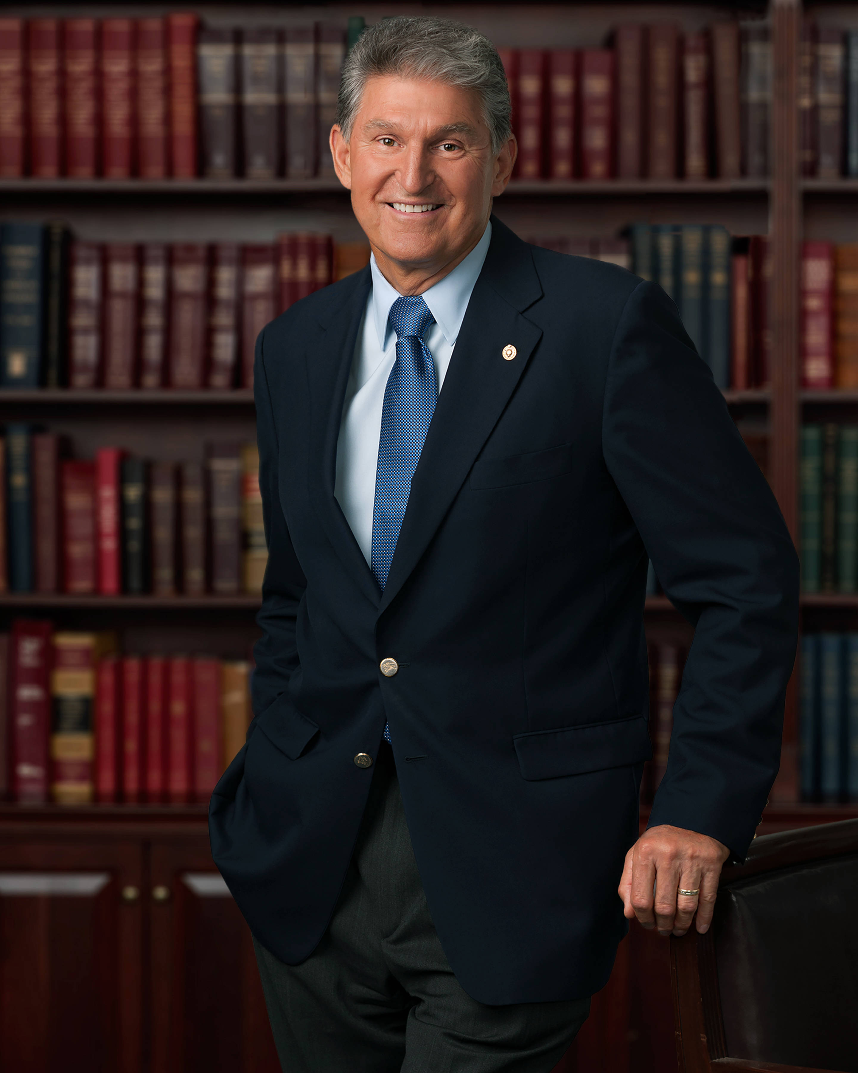 Joe Manchin 在圖書館 壁紙 — 一位微笑的男士身穿深色西裝，自信地站在充滿皮革裝訂書籍的豐富木書架背景前。