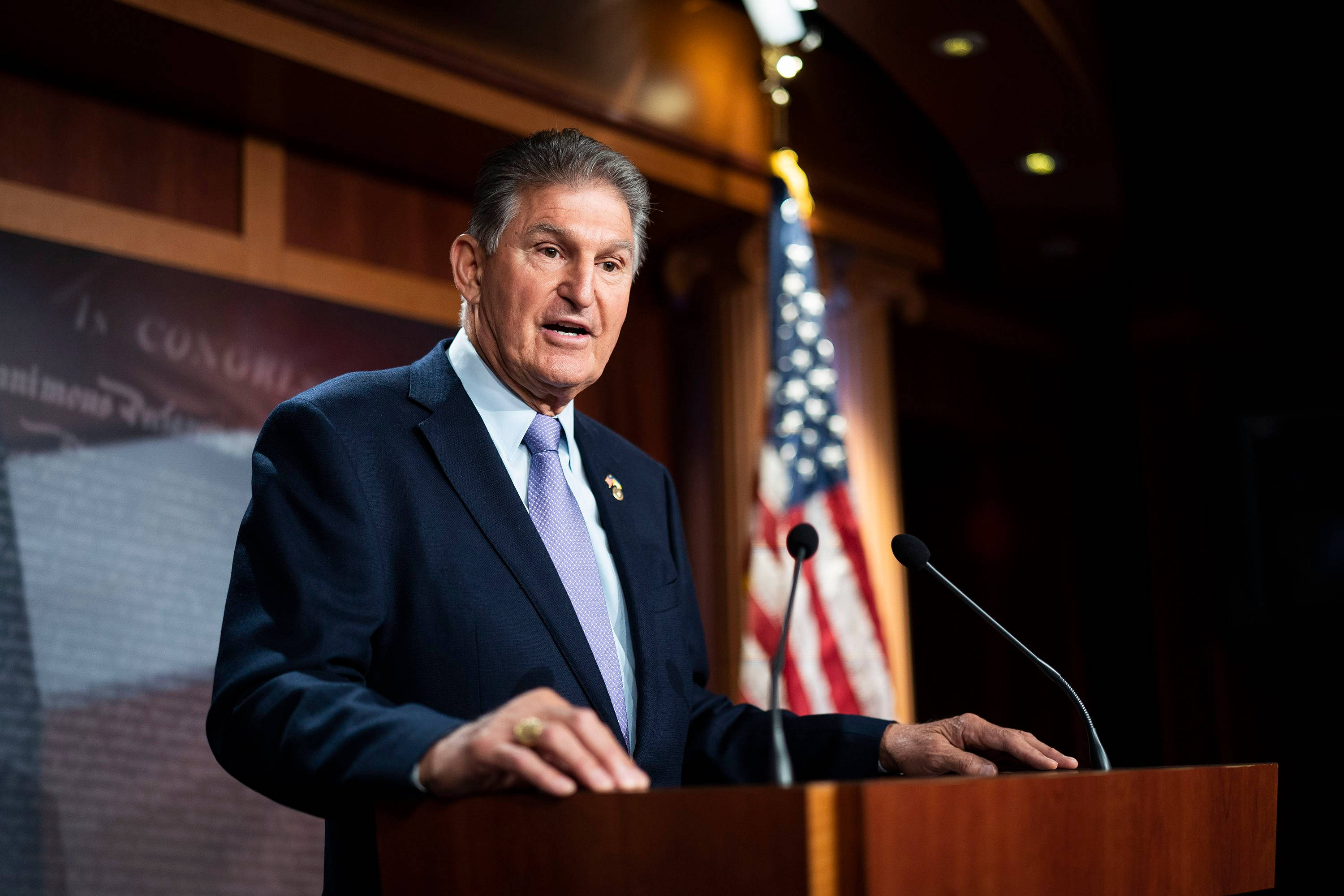 Joe Manchin 在發表講話 壁紙 — 一名身穿深色西裝的男子站在木製講台上對著麥克風講話，身後有美國國旗，在正式的室內環境中。