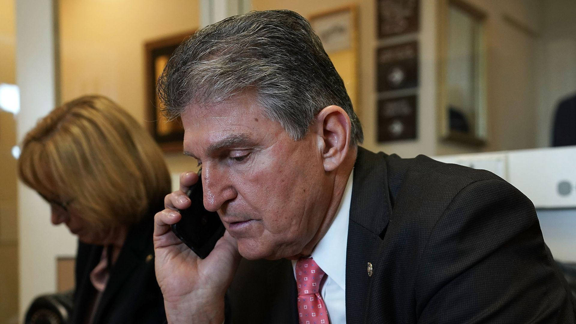 Joe Manchin 接聽電話 壁紙 — 一個嚴肅嘅男人著住西裝喺辦公室講電話，而一個女人坐喺附近。