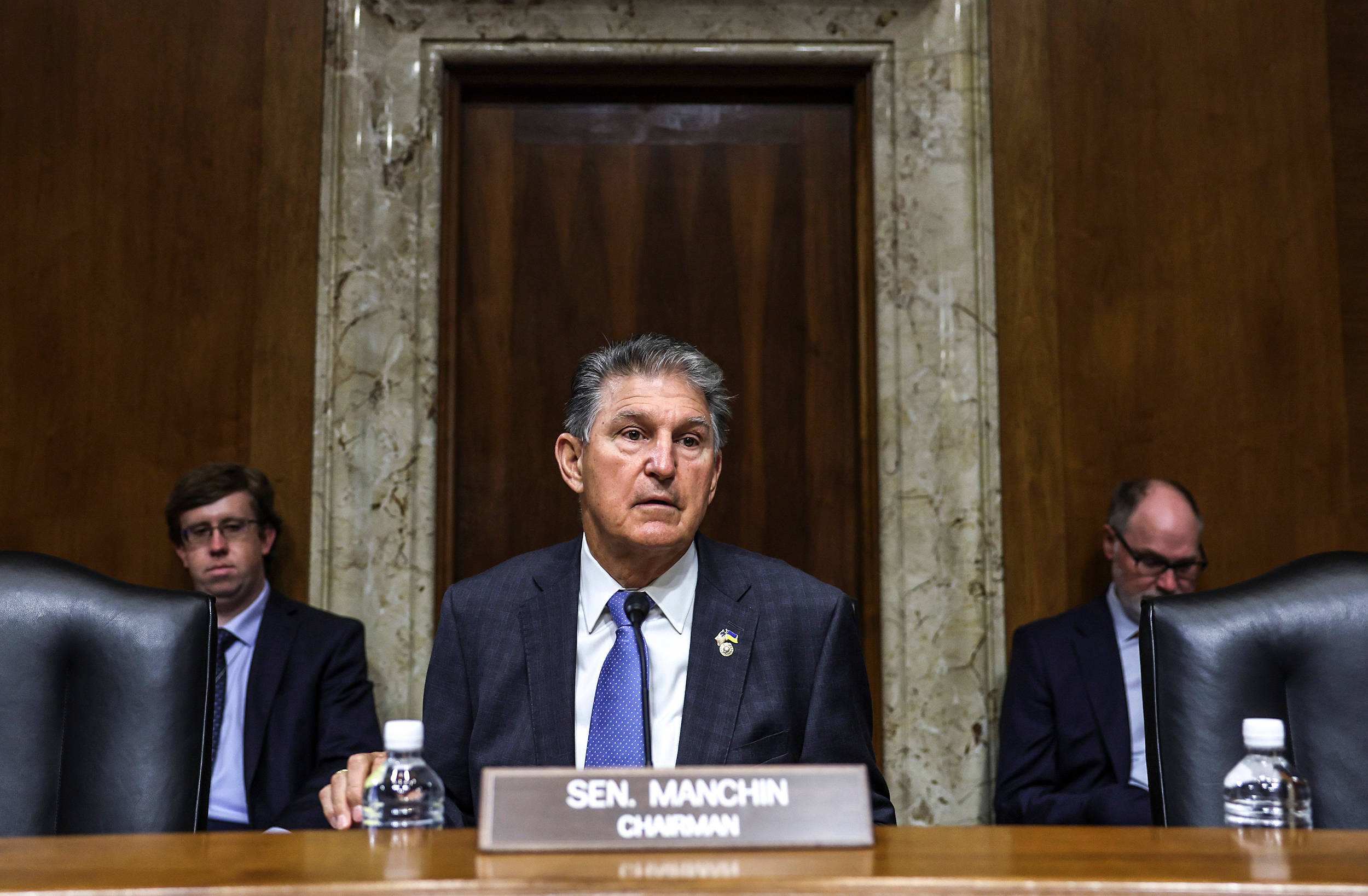 Joe Manchin 與兩名保安 壁紙 — 曼欽參議員坐在一張木製聽證會桌前，桌上放著名牌和水瓶，兩旁有兩名穿西裝的男子，身處一個有大理石和木鑲板的正式會議廳。