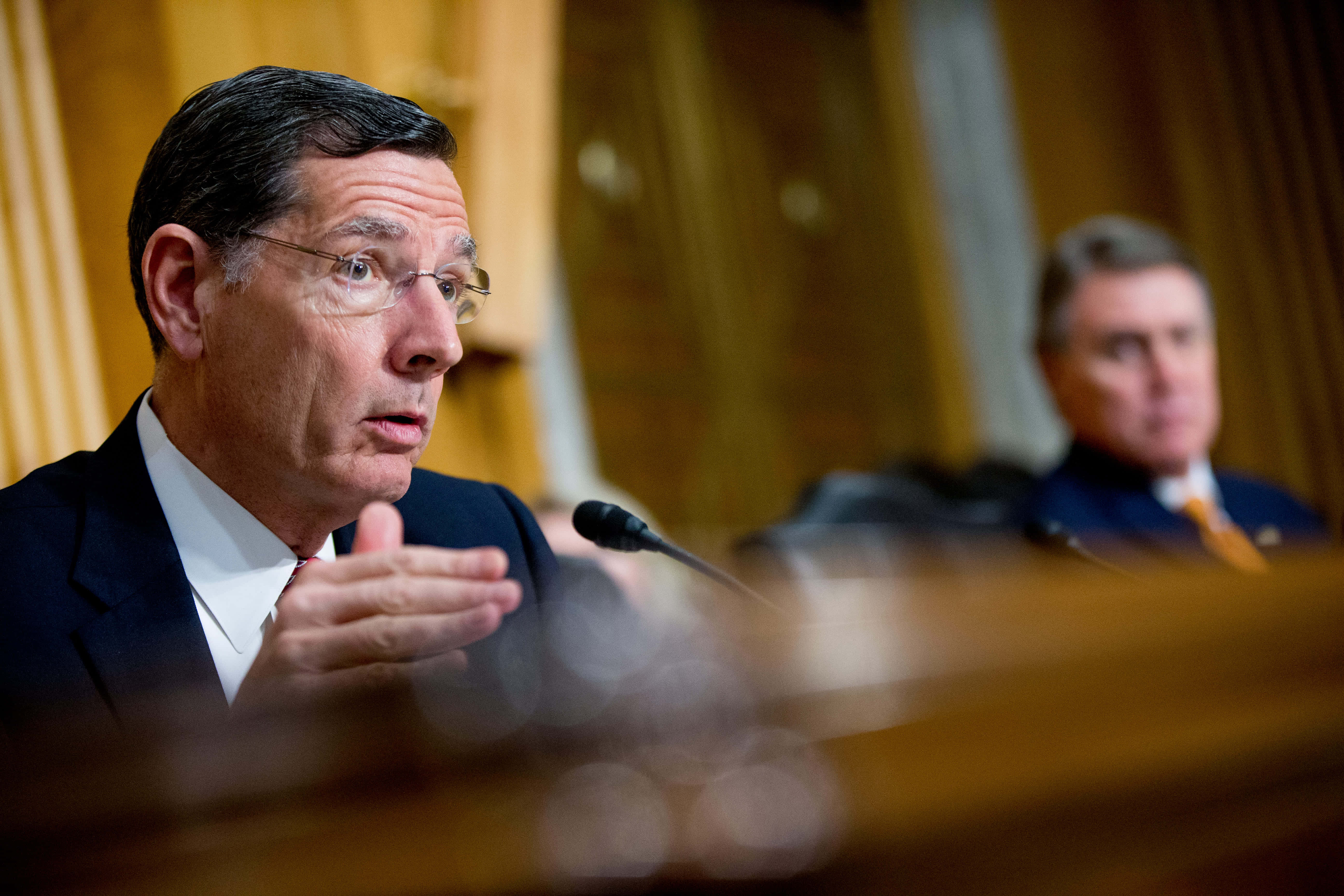 John Barrasso 在會議中發言 壁紙 — 一名身穿西裝、戴眼鏡的男子在木桌前用咪高峰認真地說話，背景中另一名男子模糊地坐着。