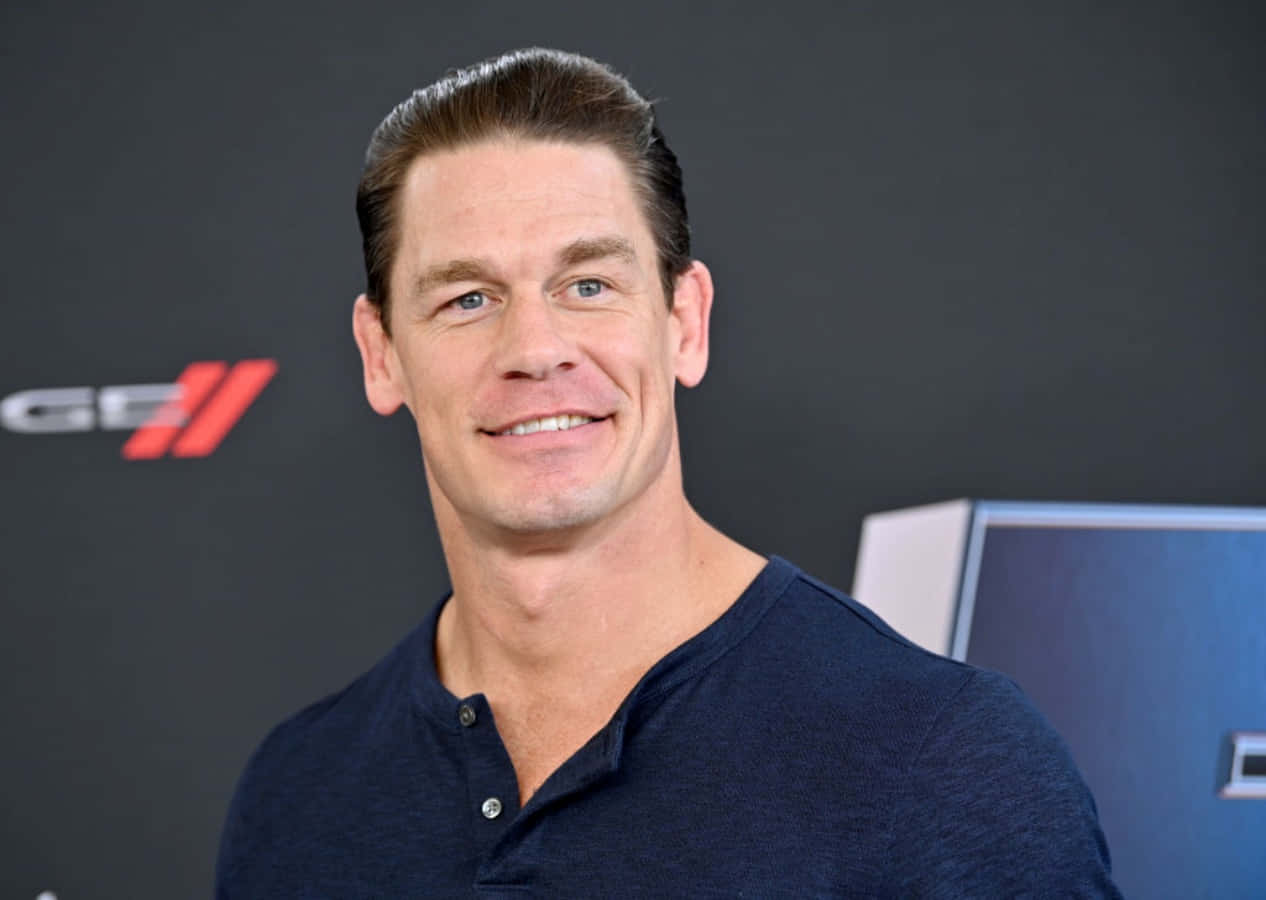 John Cena 桌布 壁紙 — John Cena 穿著深藍色亨利衫，在品牌活動背景前溫暖微笑。