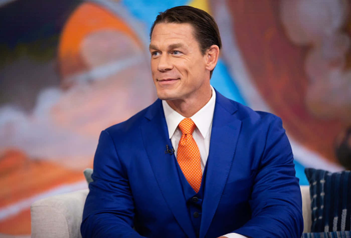 John Cena 桌布 壁紙 — John Cena 自信地坐在鮮藍色西裝和橙色領帶中，背景是色彩繽紛的模糊效果。