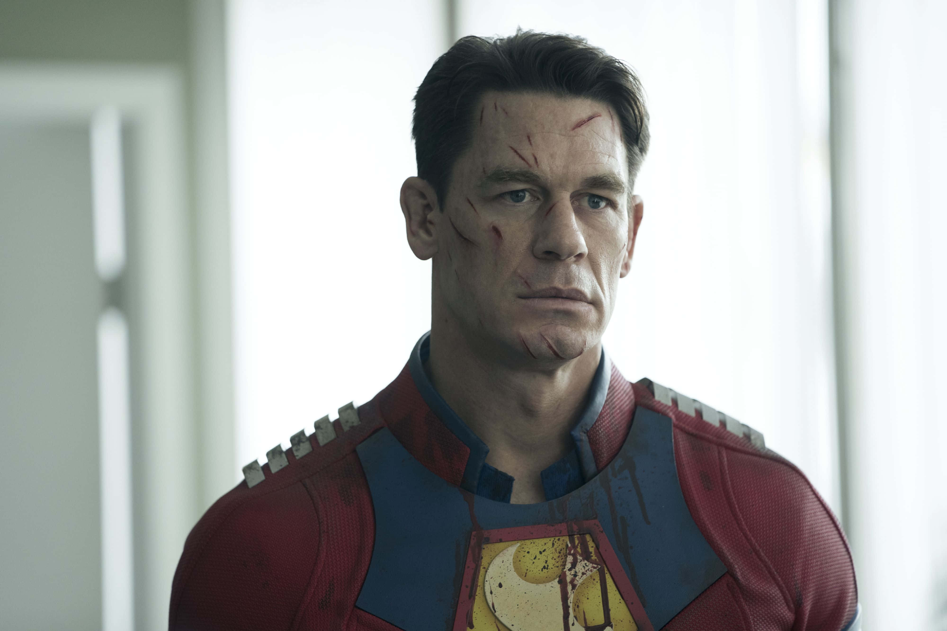 Peacemaker 桌布 壁紙 — John Cena 飾演超人，滿臉鮮血，服裝破爛，站在明亮的簡約房間裡。
