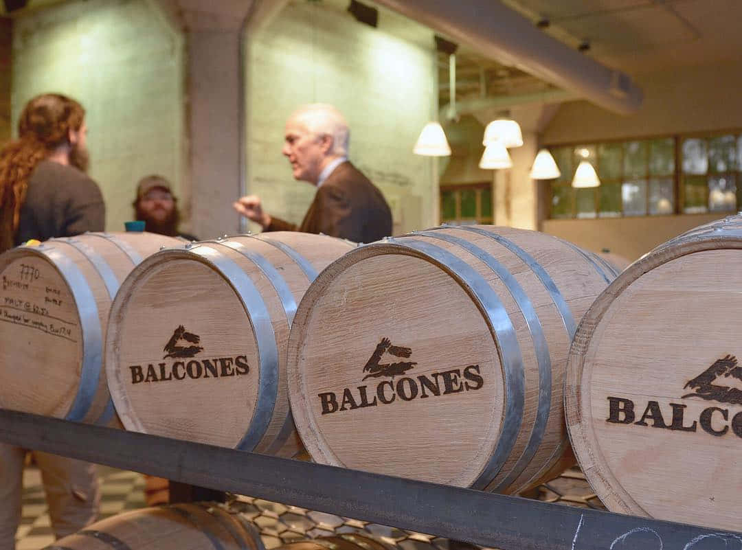 帶有 Balcones 標誌的木製威士忌桶，釀酒廠內人們在交談。