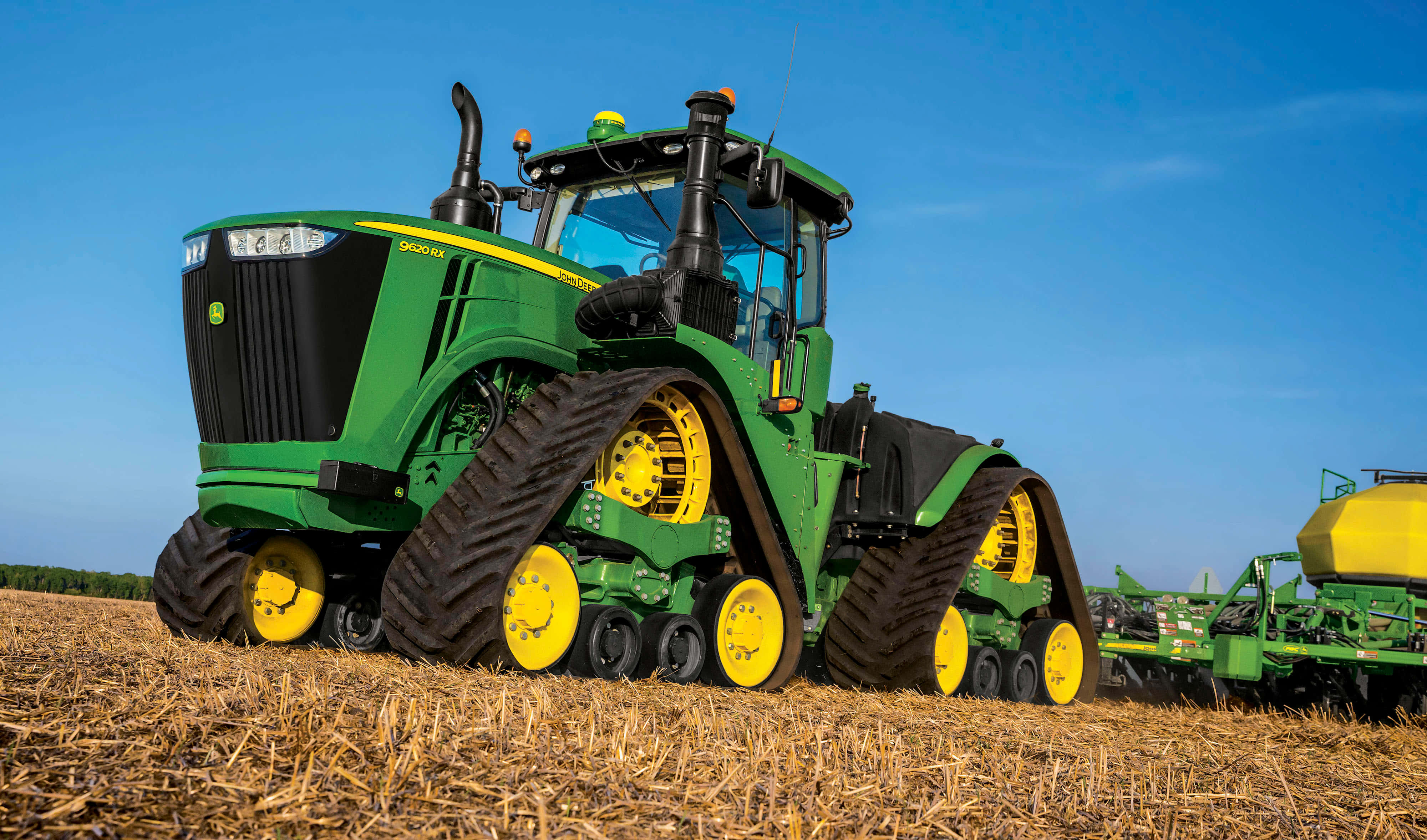 John Deere 4-Track 拖拉機圖片 壁紙 — 一輛綠色約翰迪爾履帶拖拉機帶有黃色輪子，站在晴朗藍天下已收割的田地中。