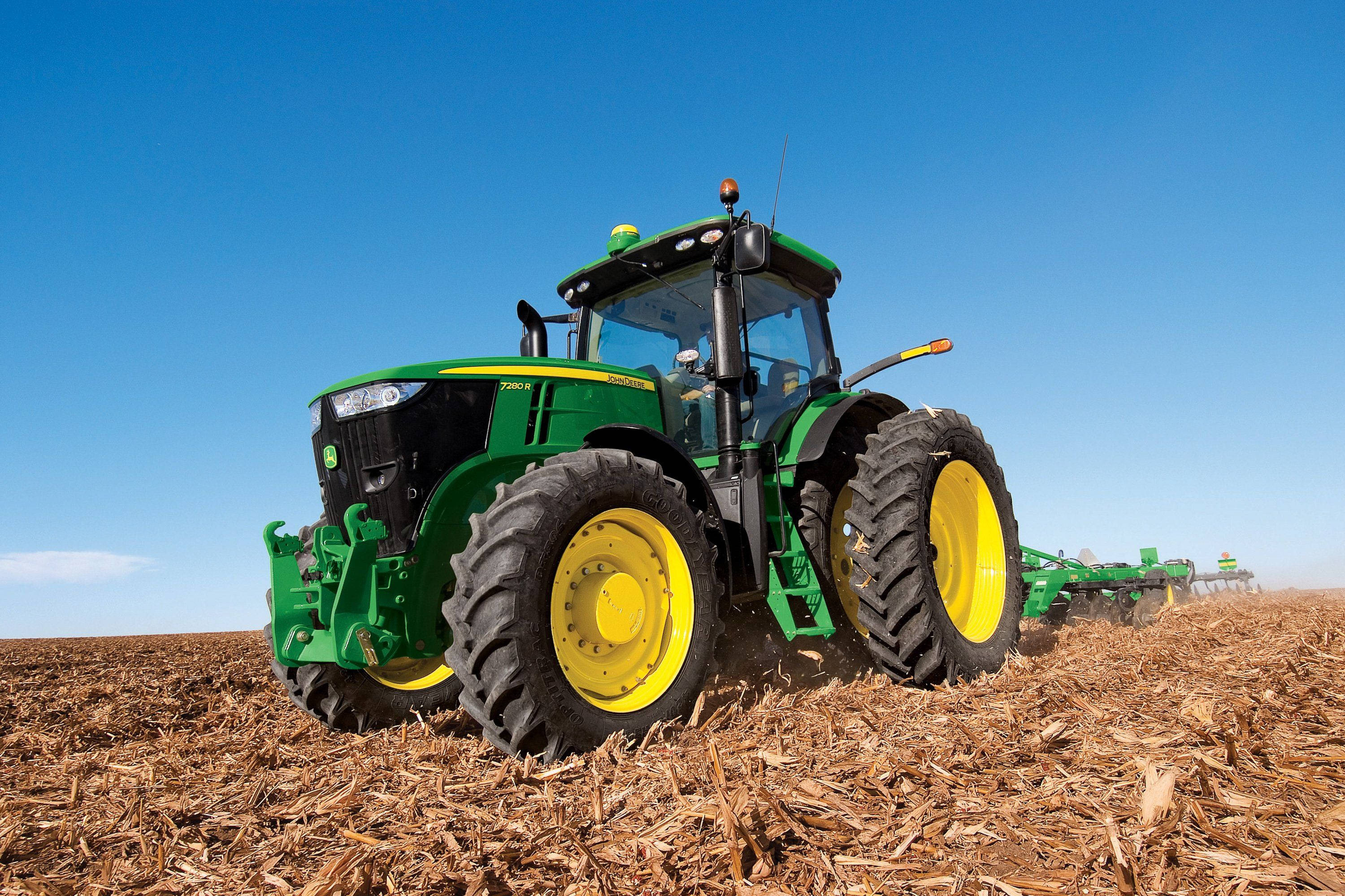 綠色John Deere 7R拖拉機田間作業 壁紙 — 一輛綠色和黃色的約翰迪爾拖拉機在晴朗的藍天下犁過一片乾莖田地。