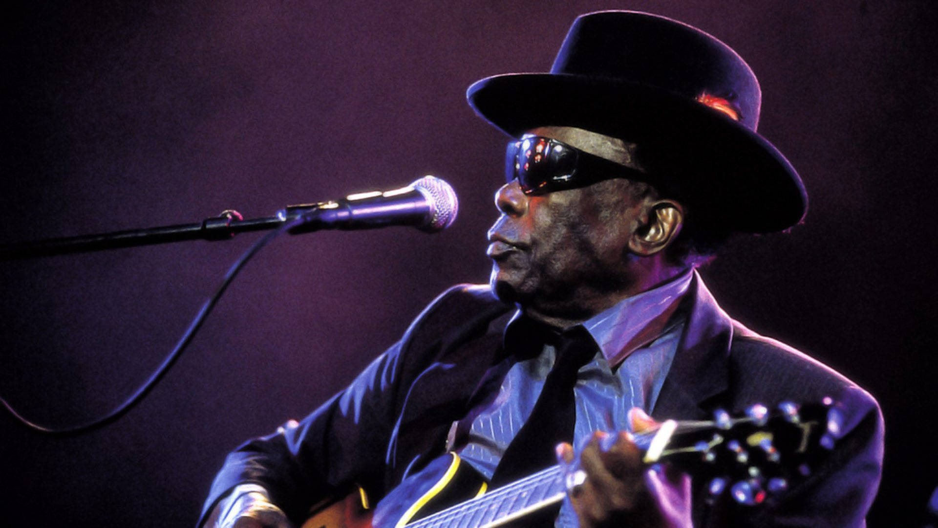 John Lee Hooker 感人演唱照 壁紙 — 一位戴黑色帽子和太陽眼鏡的音樂家在紫色舞台燈光下彈結他，並對著麥克風唱歌。