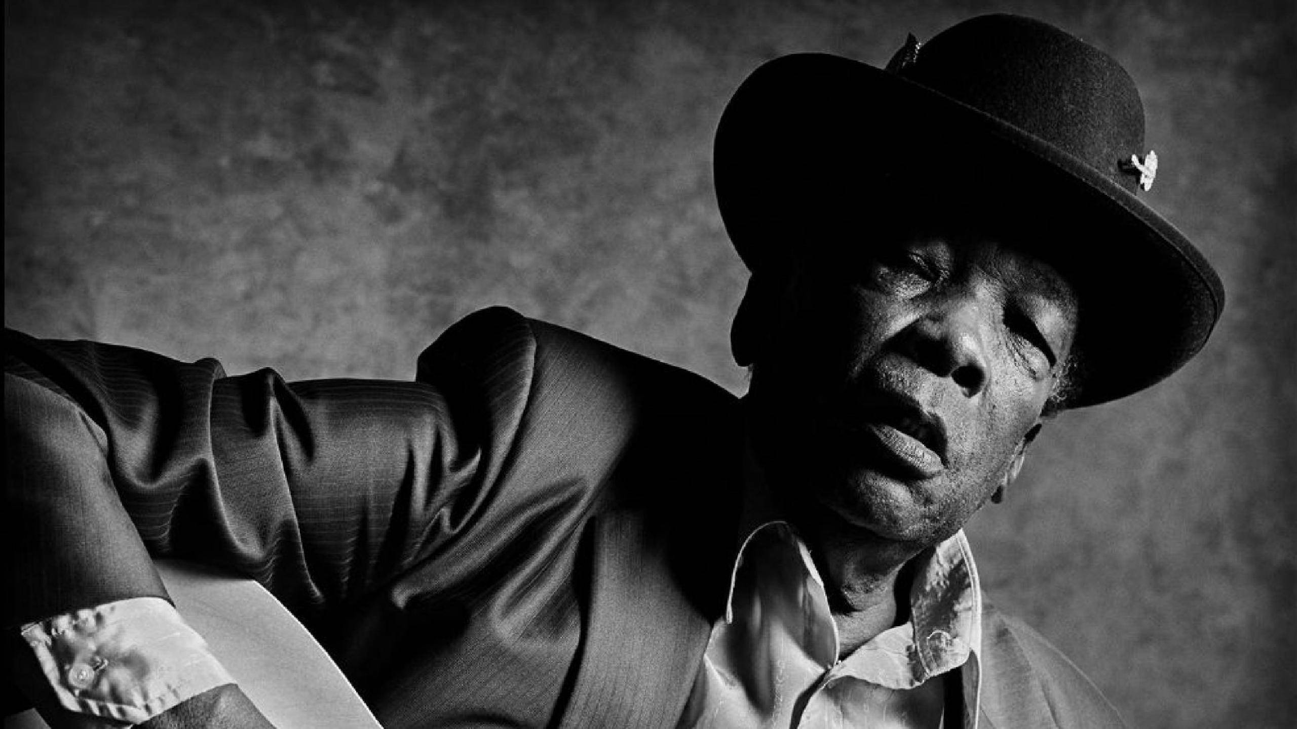 John Lee Hooker 單色照 壁紙 — 一個戴著紳士帽和西裝的男人，低頭帶著強烈表情的黑白肖像。