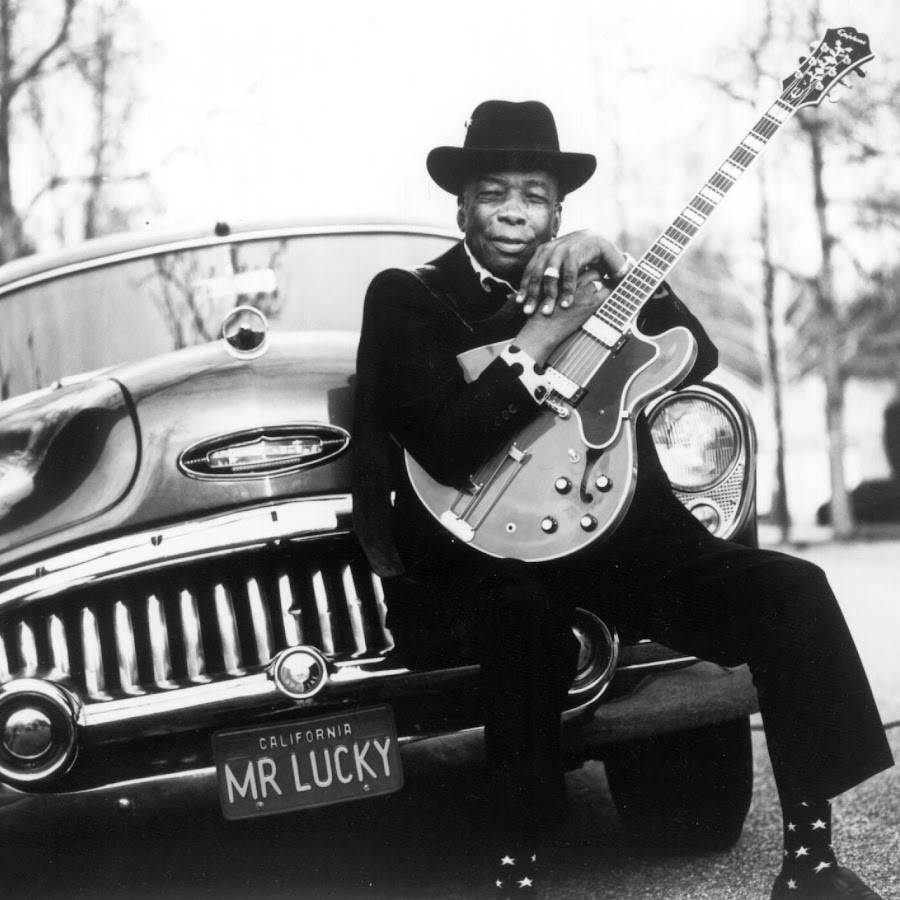 John Lee Hooker 在經典汽車前拍攝 壁紙 — 一個黑白肖像，戴著費多拉帽的男人坐在一輛帶有'MR LUCKY'車牌的復古汽車引擎蓋上，手持結他。