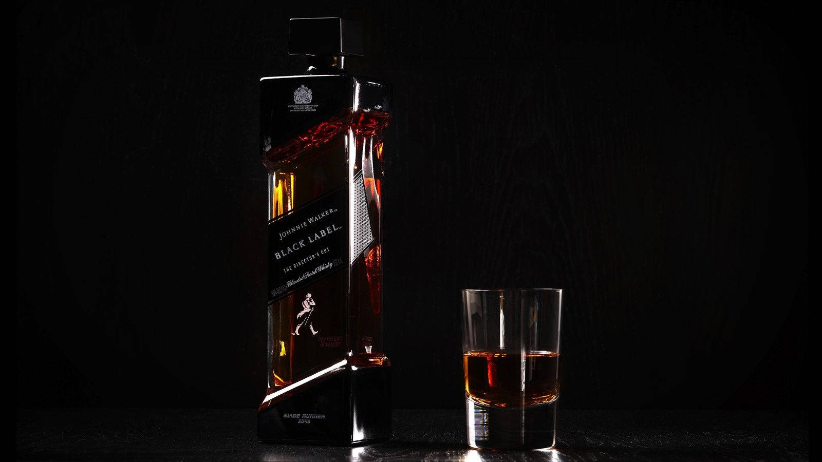 Johnnie Walker黑牌威士忌與威士忌量杯的時尚拍攝 壁紙 — 在深色紋理背景前，一瓶纖細的黑色尊尼獲加黑牌威士忌旁邊放著一杯琥珀色液體。