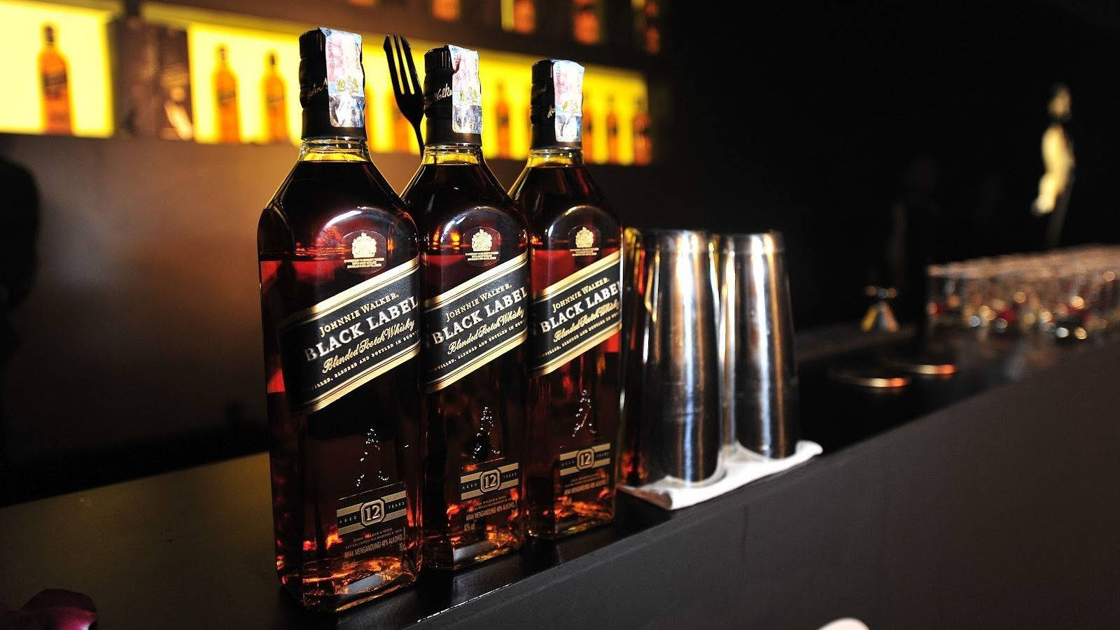 吧檯上的三瓶Johnnie Walker黑牌威士忌 壁紙 — 三瓶尊尼獲加黑牌威士忌在溫暖背光下閃耀，放置在時尚的吧檯上，旁邊有雞尾酒調酒器。