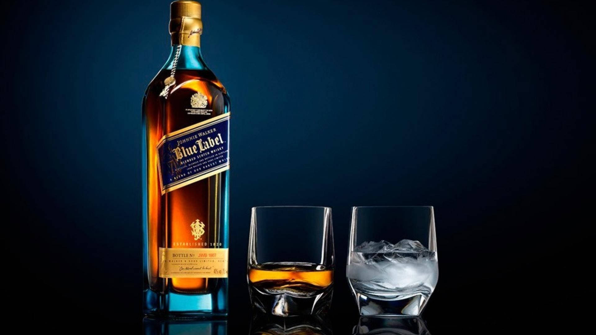 Johnnie Walker威士忌瓶身的奢華典雅風貌 壁紙 — 一個時尚的Johnnie Walker Blue Label威士忌酒瓶旁邊有兩個玻璃杯，一個裝滿琥珀色液體，另一個裝有冰塊，背景是深藍色漸變。