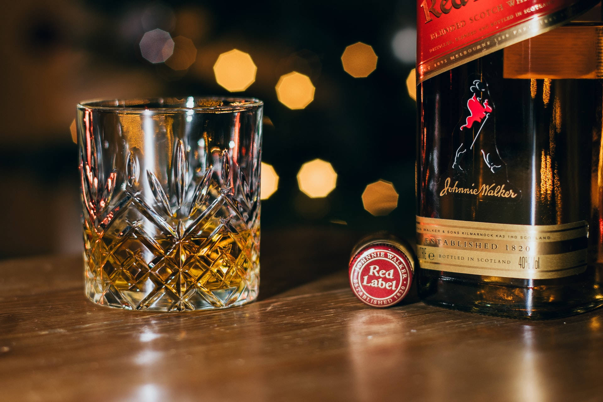 Johnnie Walker紅牌威士忌與木桌上的威士忌杯 壁紙 — 一個裝有琥珀色威士忌的水晶杯放在約翰走路紅牌酒瓶和瓶蓋旁邊，置於木質吧檯上，背景有溫暖的散景燈光。