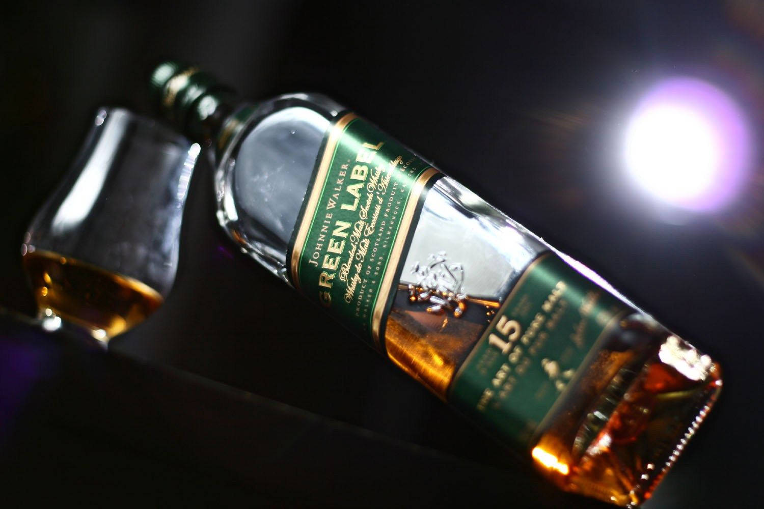 Johnnie Walker綠牌威士忌倒入風暴杯 壁紙 — 一瓶傾斜的Johnnie Walker Green Label威士忌，旁邊放著一個玻璃杯，在黑暗背景下被明亮的紫色鏡頭光暈照亮。