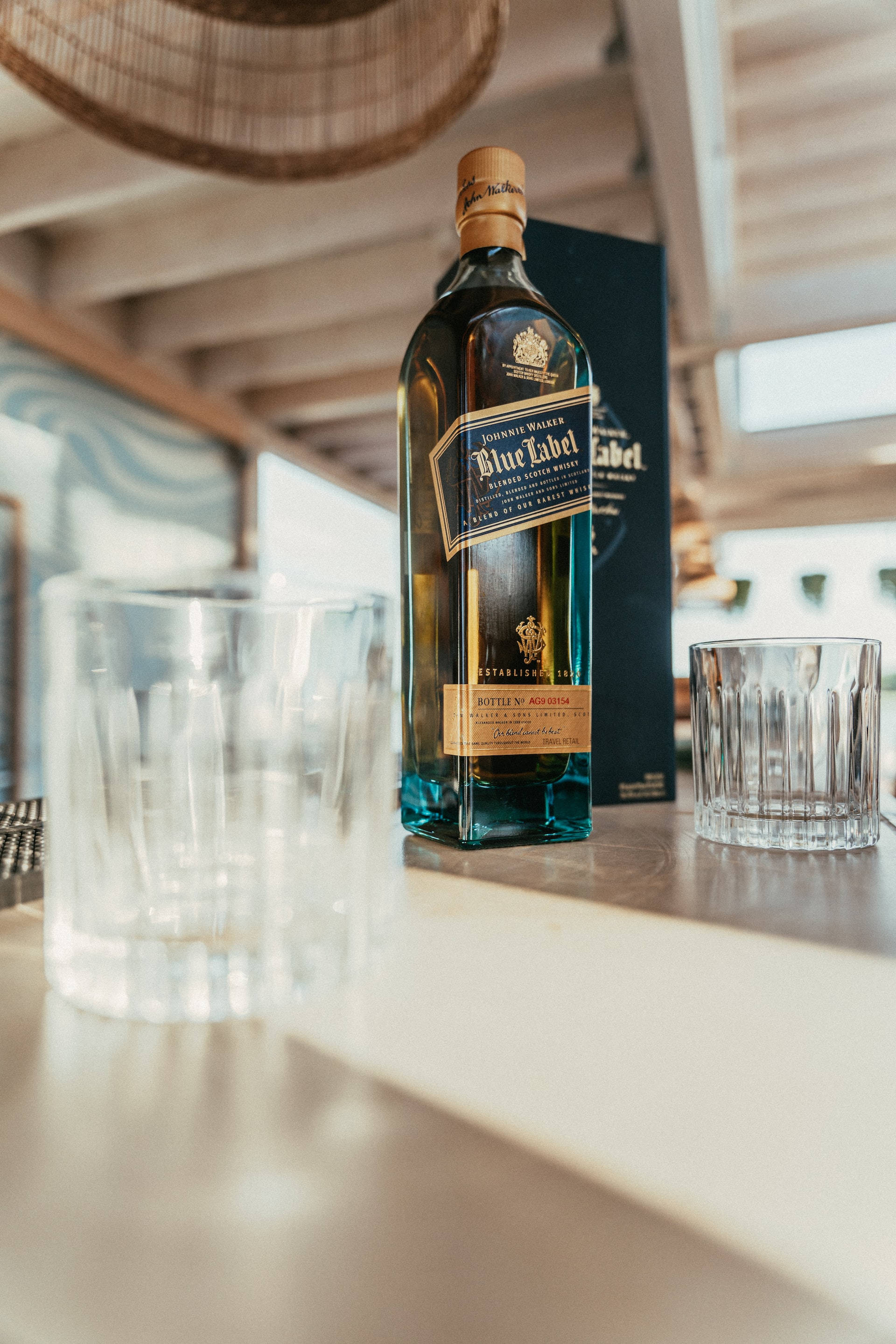 Johnnie Walker藍牌瓶身於酒杯間的吸睛拍攝 壁紙 — 一瓶尊尼獲加藍牌威士忌優雅地放在陽光照射的木製吧檯上，旁邊有兩個水晶杯。