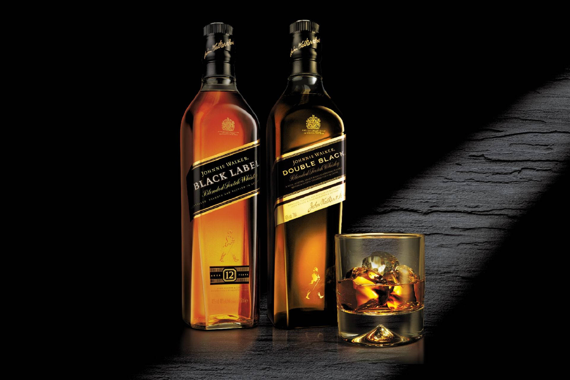 Johnnie Walker蘇格蘭威士忌倒入經典低球杯 壁紙 — 兩瓶Johnnie Walker威士忌和一杯加冰的玻璃杯放在黑暗有紋理的表面上，在戲劇性的燈光下。