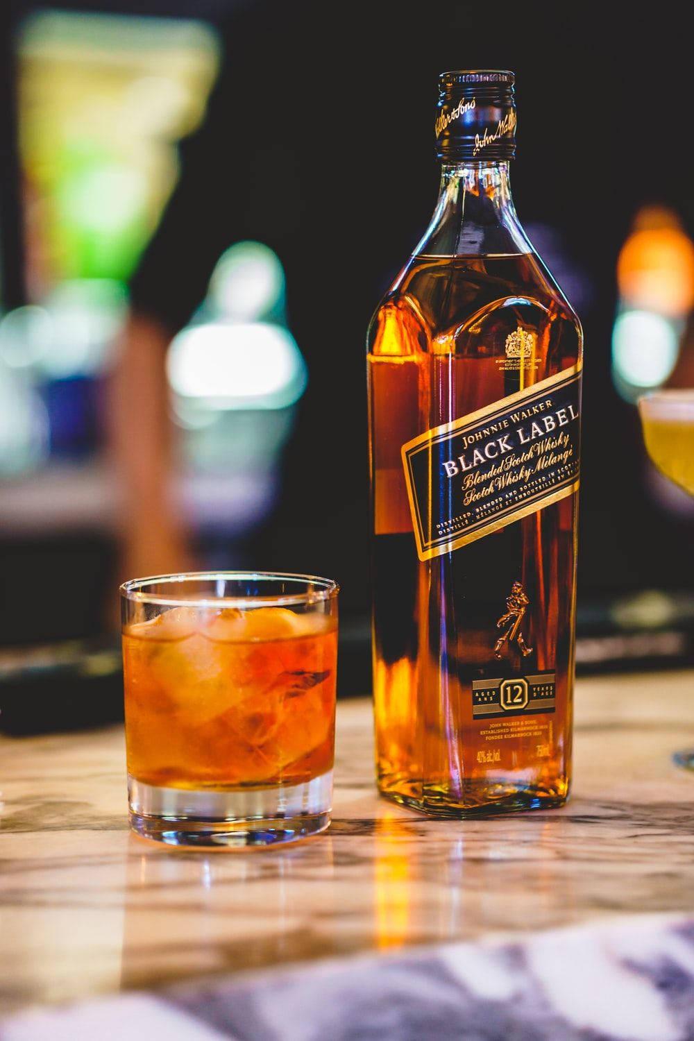 Johnnie Walker黑牌威士忌與酒杯於大理石檯面 壁紙 — 一瓶Johnnie Walker Black Label威士忌和一杯加冰的威士忌放在大理石吧檯上，背景是模糊的霓虹燈光。