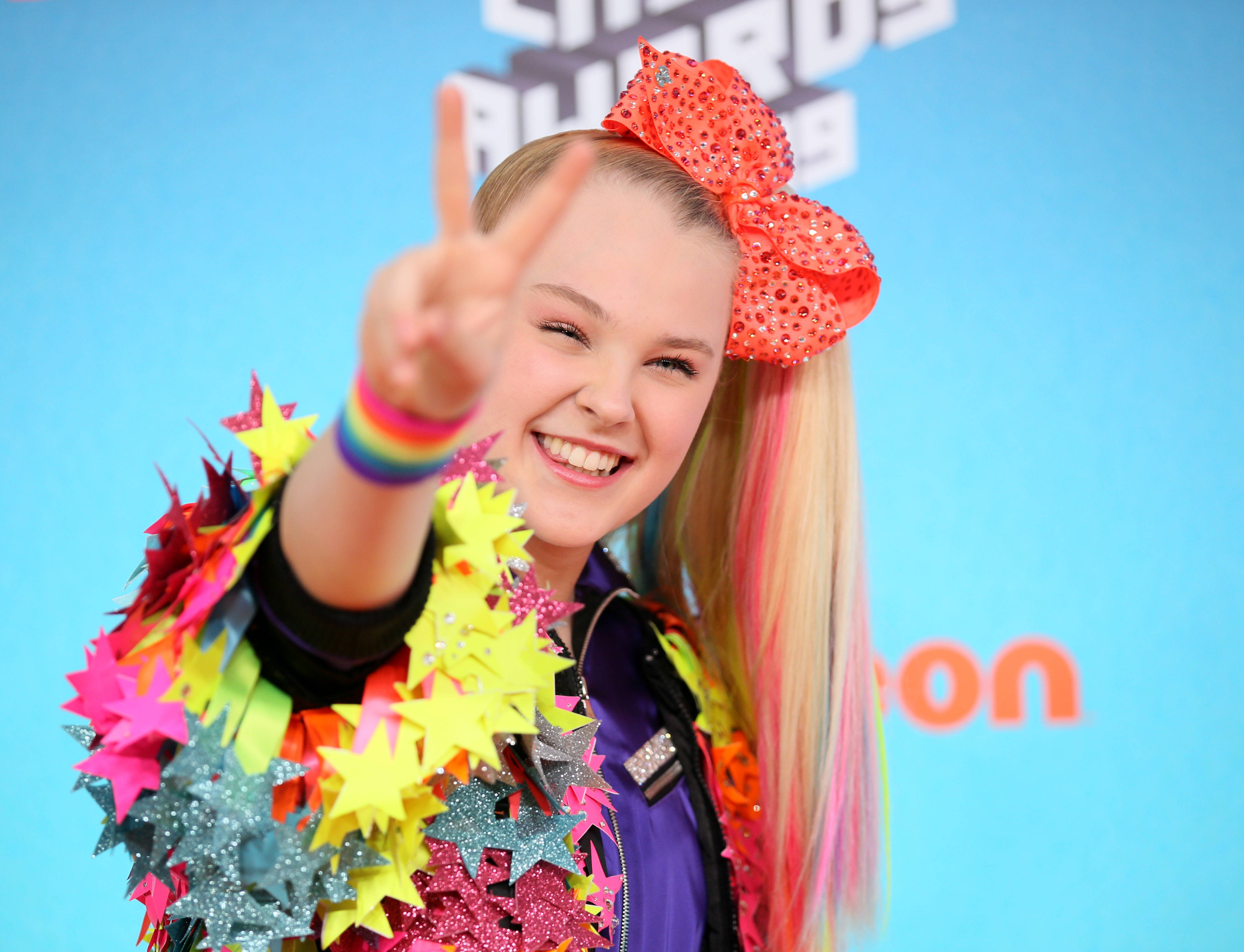 Jojo Siwa 做 V 字手勢 壁紙 — 一位開朗的小女孩戴着一個巨大的閃亮紅色蝴蝶結，對着鏡頭露出燦爛的笑容並比出和平手勢。