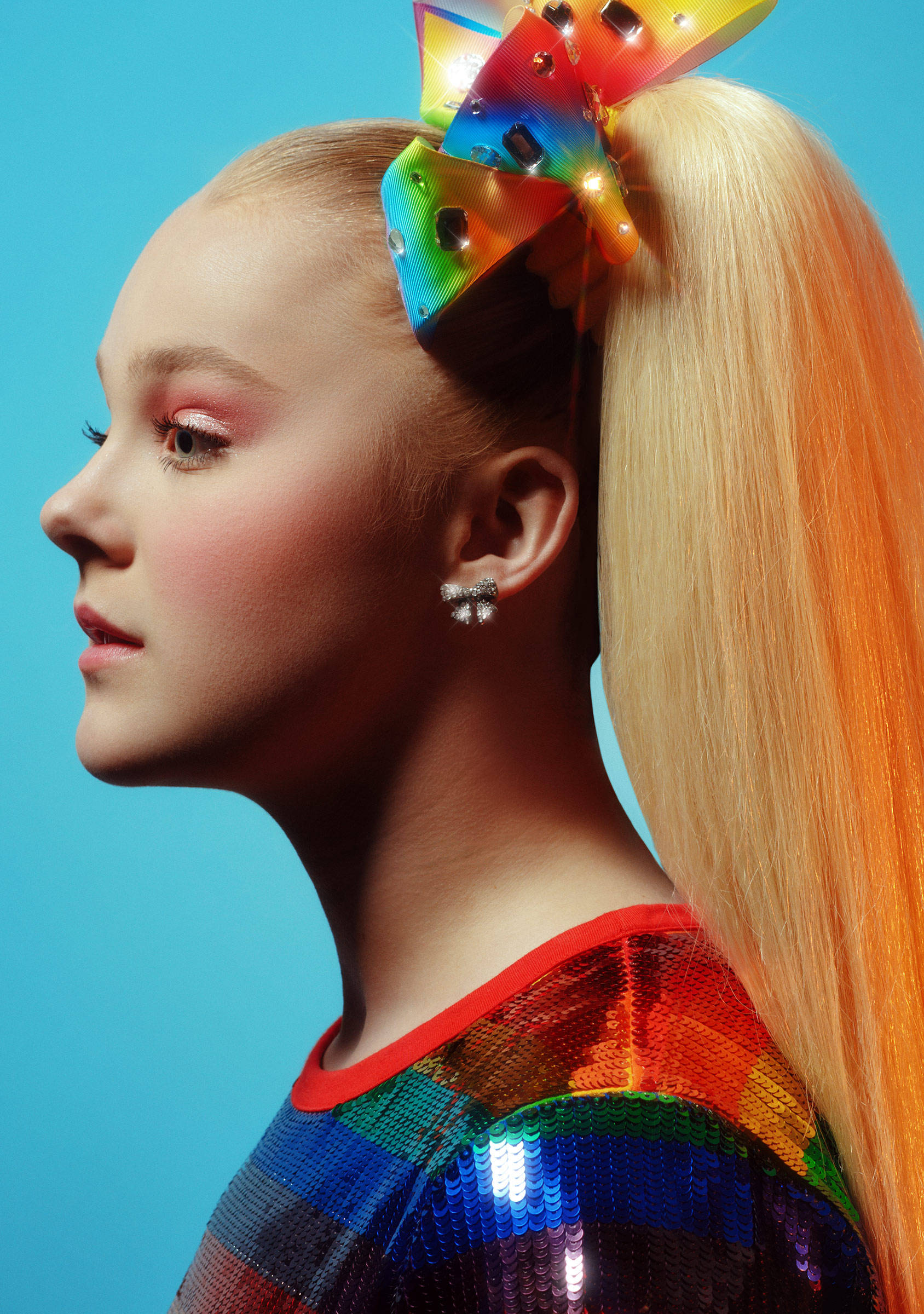 Jojo Siwa 側面剪影 壁紙 — 一位年輕女性紮著金色高馬尾，戴著閃閃發光的彩虹蝴蝶結，穿著亮片上衣，背景為純藍色。