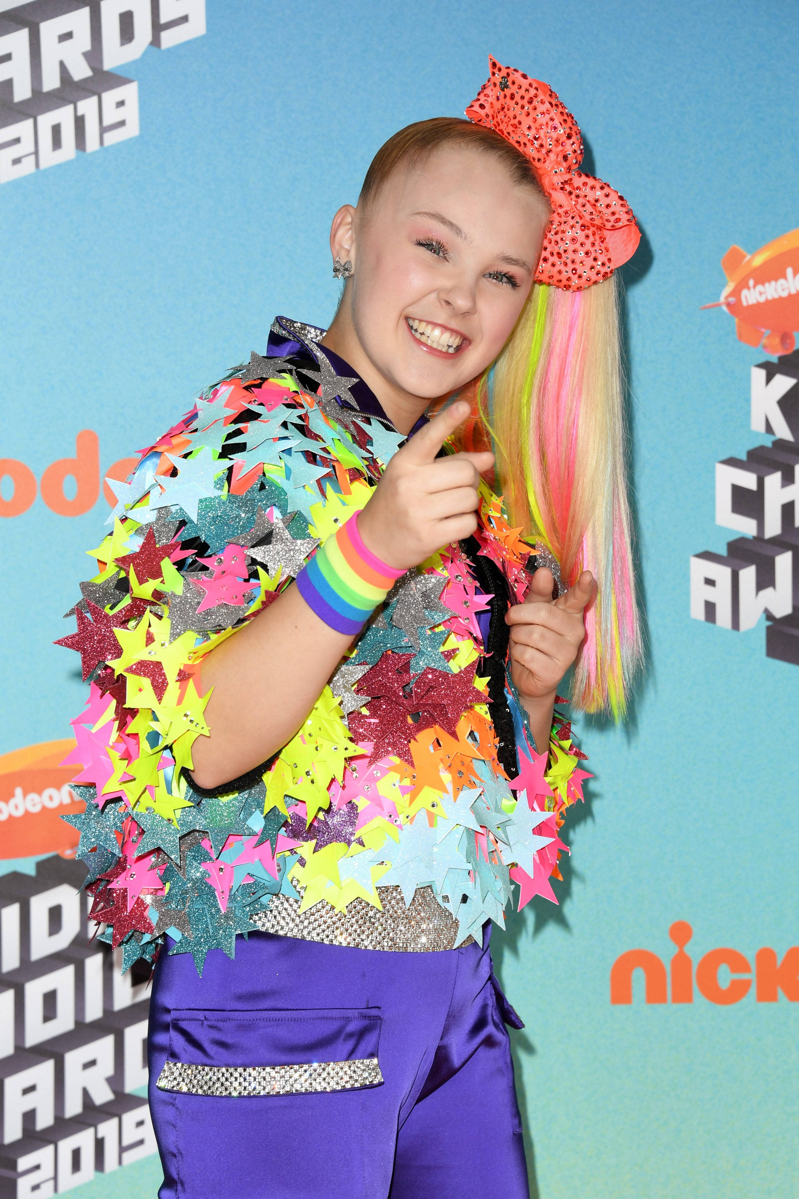 Jojo Siwa 出席尼克兒童選擇獎 壁紙 — 一個微笑的年輕女孩，彩虹頭髮和一個巨大的蝴蝶結，穿著閃閃發光的星星外套出席2019年兒童選擇獎。