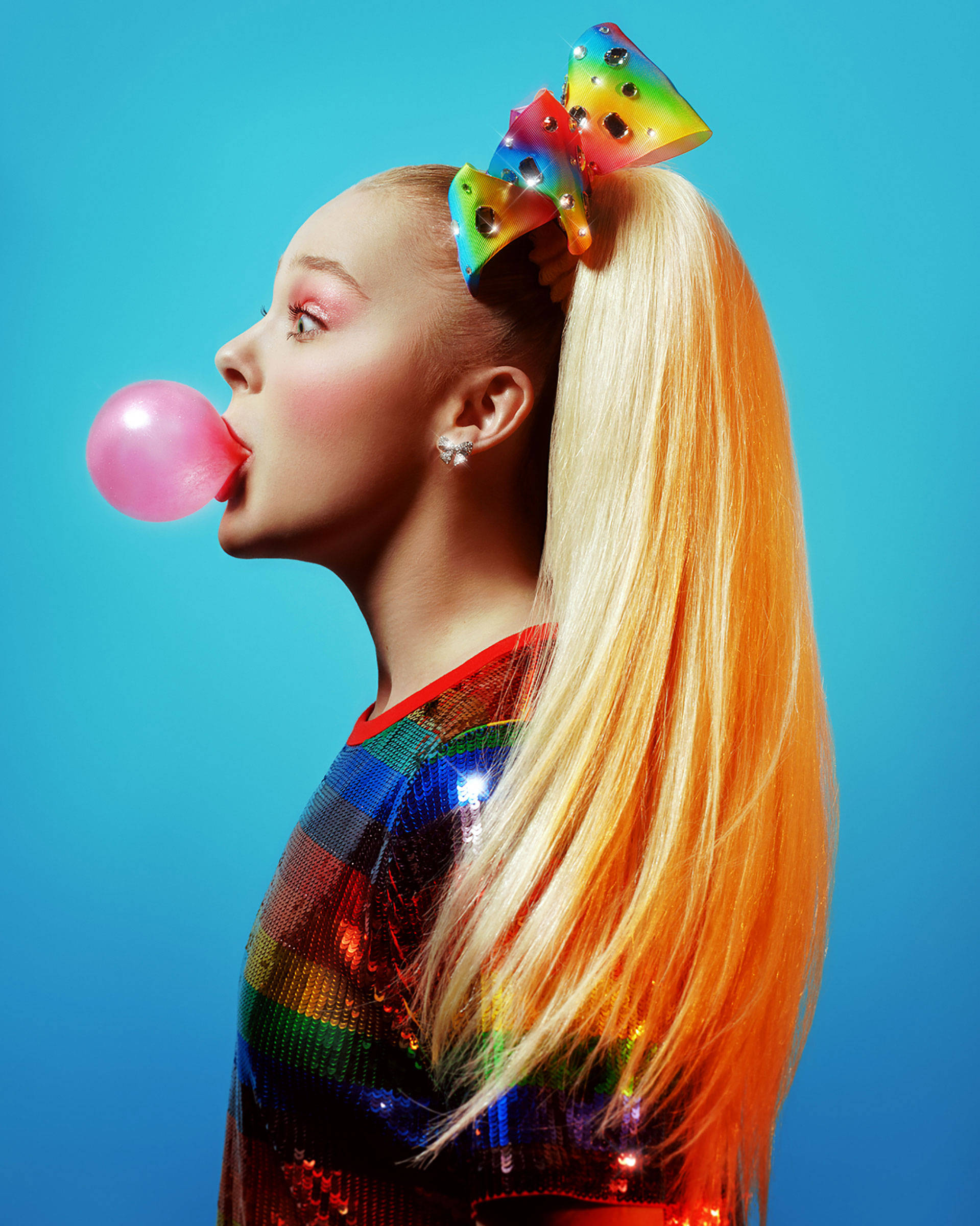 Jojo Siwa 吹泡泡糖 壁紙 — 一位金髮年輕女子戴著彩虹蝴蝶結，在鮮豔的藍色背景下吹出粉紅色泡泡糖泡泡。
