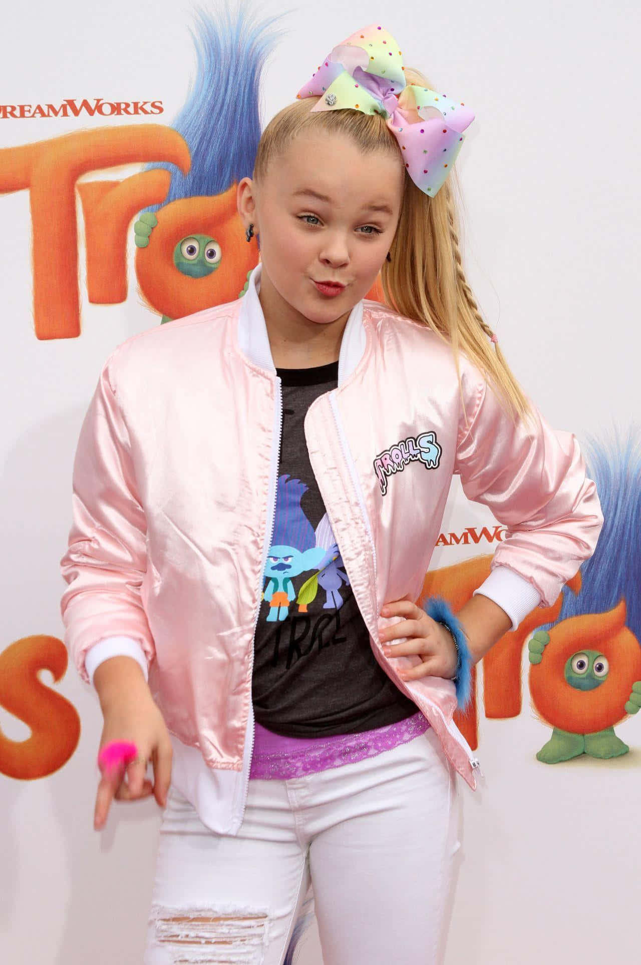 jojo siwa 桌布 壁紙 — JoJo Siwa 在一個有色彩繽紛角色背景的 Trolls 活動中，自信地穿著粉紅色 bomber 外套和白色破洞牛仔褲擺姿勢。