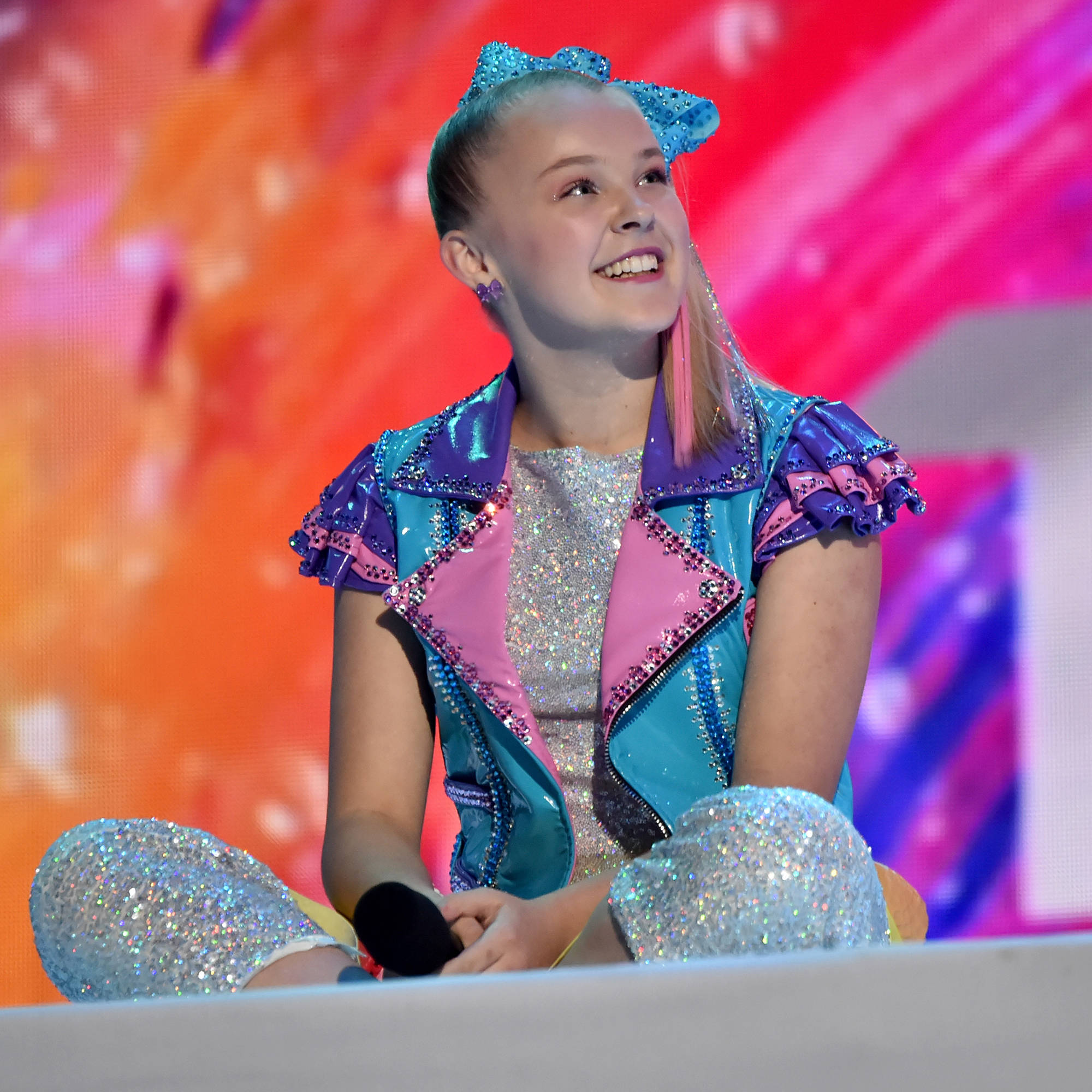 Jojo Siwa 坐姿照 壁紙 — 一個年輕女孩穿著閃閃發光的藍色、粉紅色和銀色服裝，坐在色彩繽紛的抽象背景舞台上，露出燦爛的笑容。