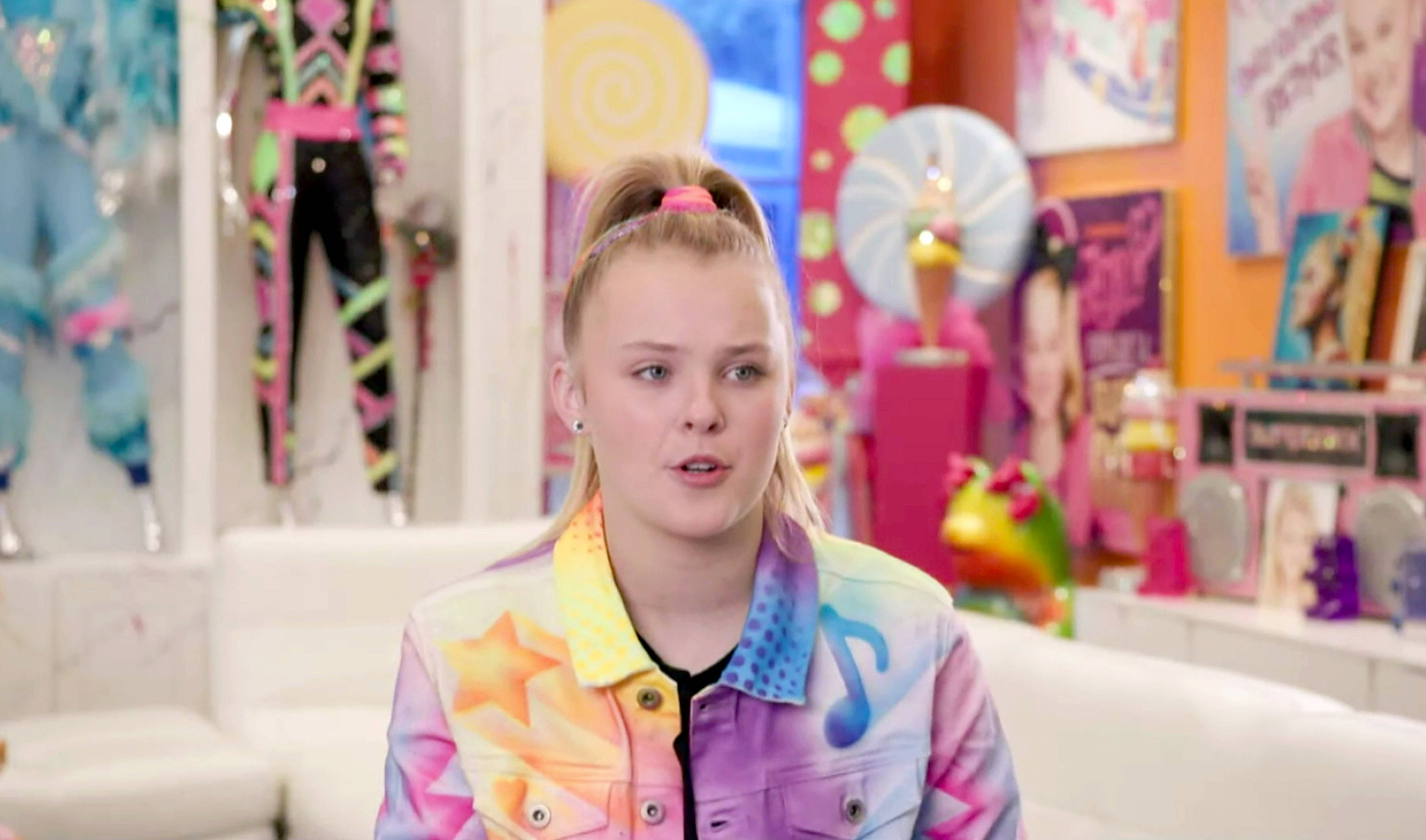 Jojo Siwa 家中訪問照 壁紙 — 一位年輕女性，金色頭髮紮成雙馬尾，身穿帶有星星和音符的彩色外套，身處充滿活力的糖果主題房間。
