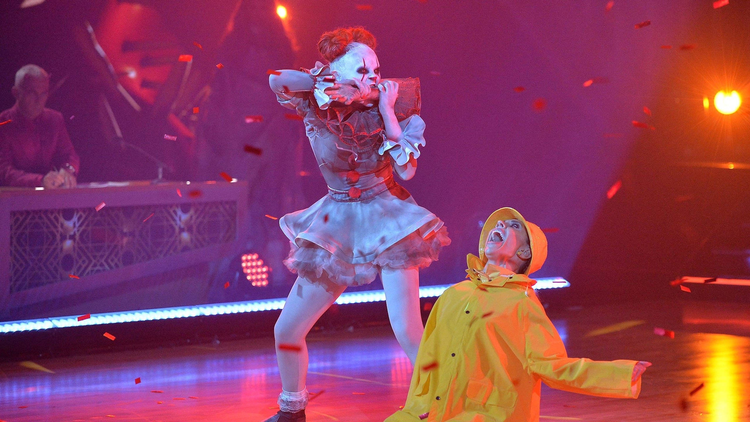 Jojo Siwa 擔任 Pennywise 角色 壁紙 — 一位表演者戴著白色骷髏面具和荷葉邊連衣裙，與另一位穿著黃色雨衣的表演者在充滿活力的舞台上對峙，五彩紙屑飄落。