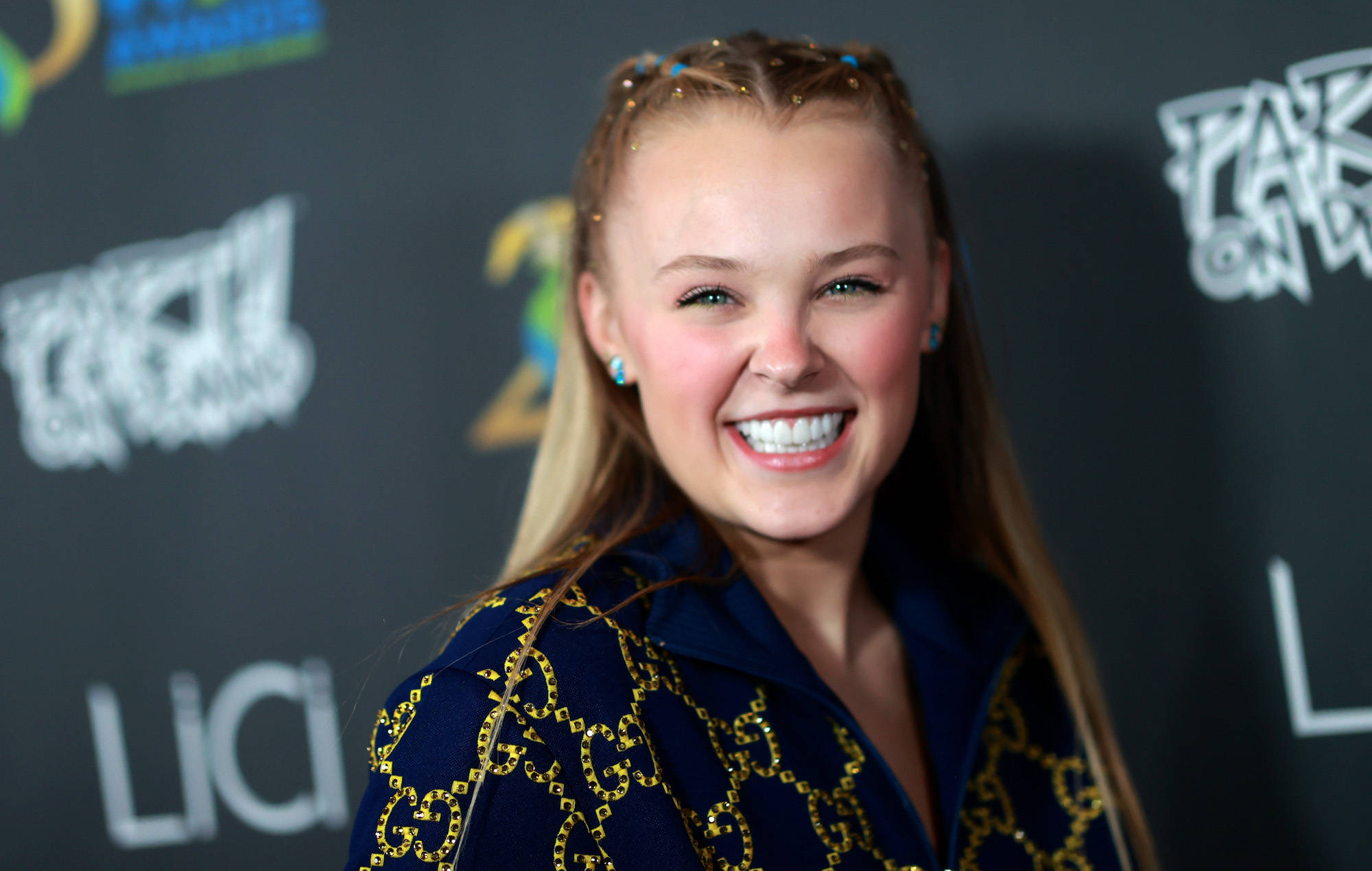 Jojo Siwa 棕色編髮造型 壁紙 — 一位微笑的年輕女性，金髮藍耳環，身穿海軍藍外套，帶有金色鏈條刺繡，在紅地毯活動中。