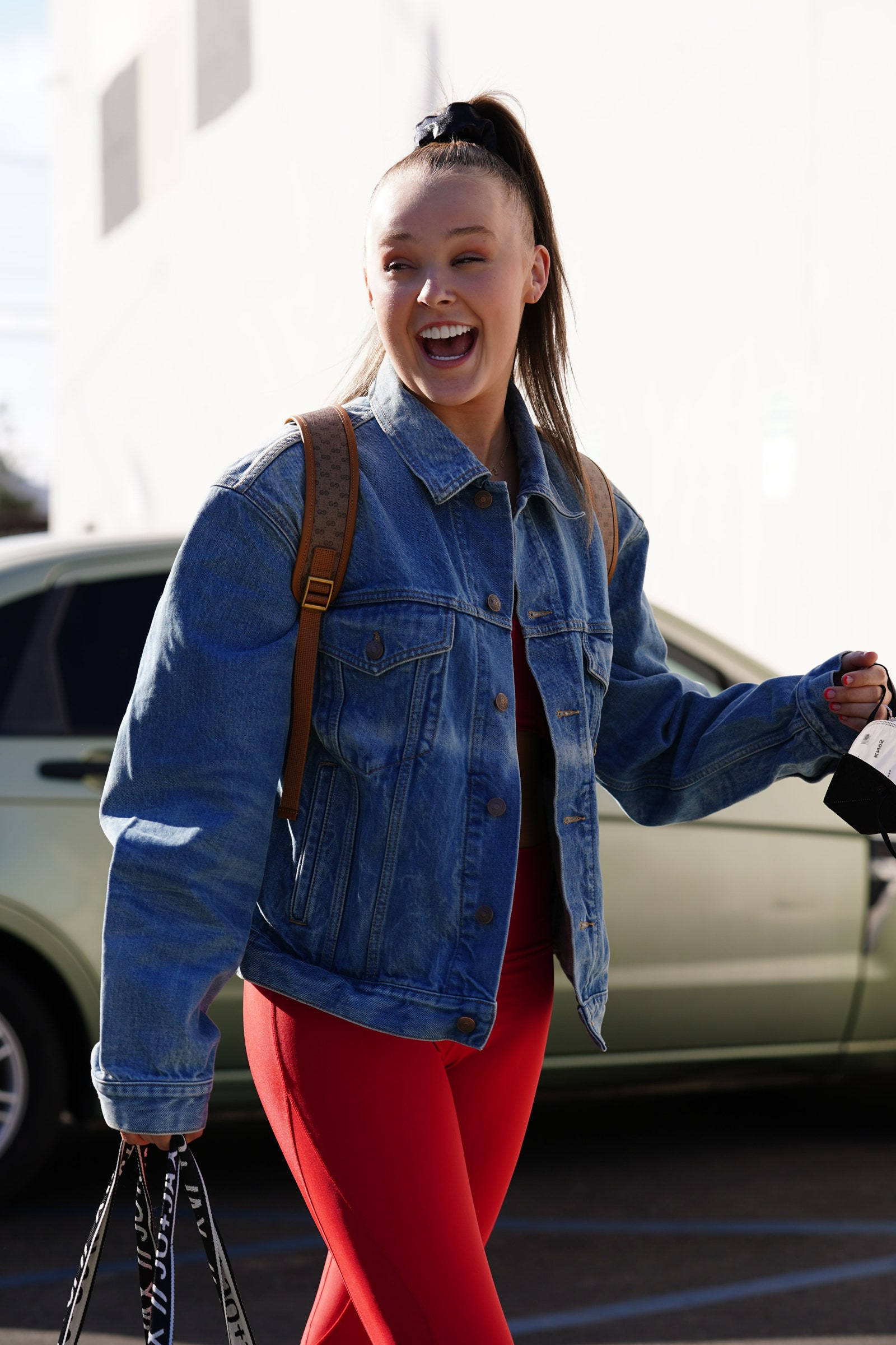 Jojo Siwa 棕髮造型 壁紙 — 一位身穿牛仔外套和紅色褲子的年輕女子，帶著購物袋在戶外快樂地笑著走路。